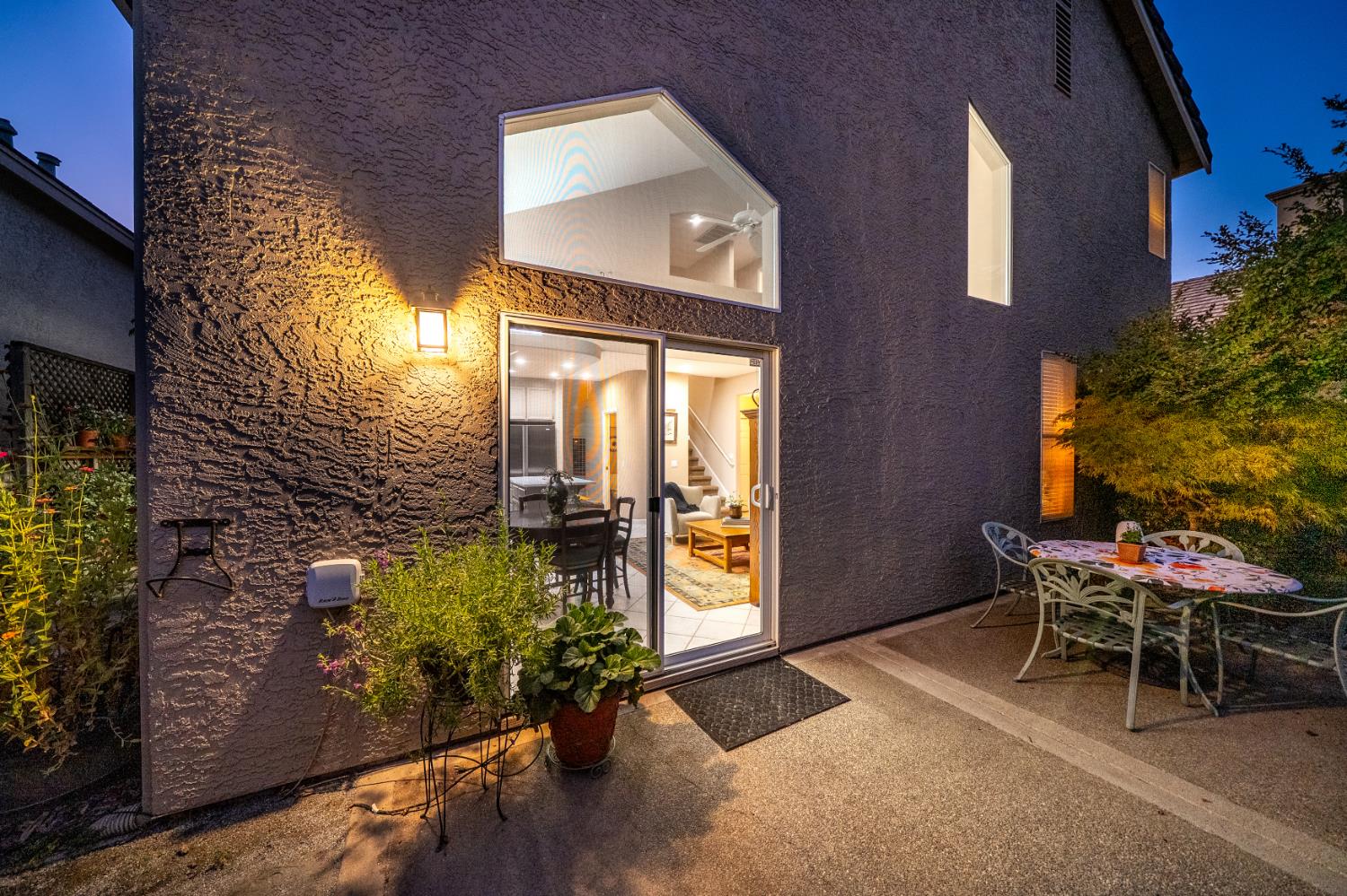 Two-story stucco home at twilight, with a warmly lit patio and furnished interior visible through sliding glass doors.