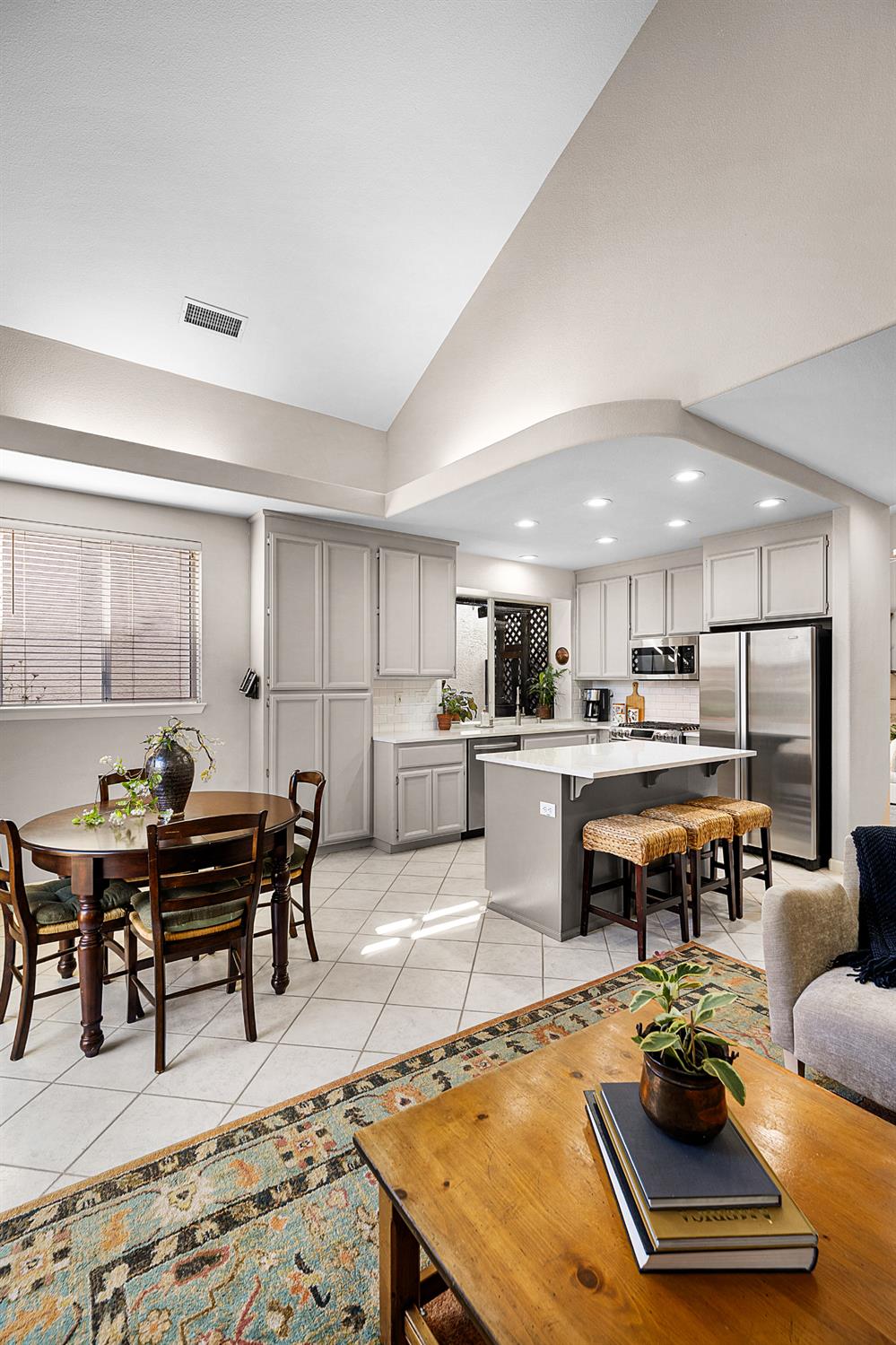 Open kitchen and dining area with a gray island, woven stools, and vaulted ceiling.