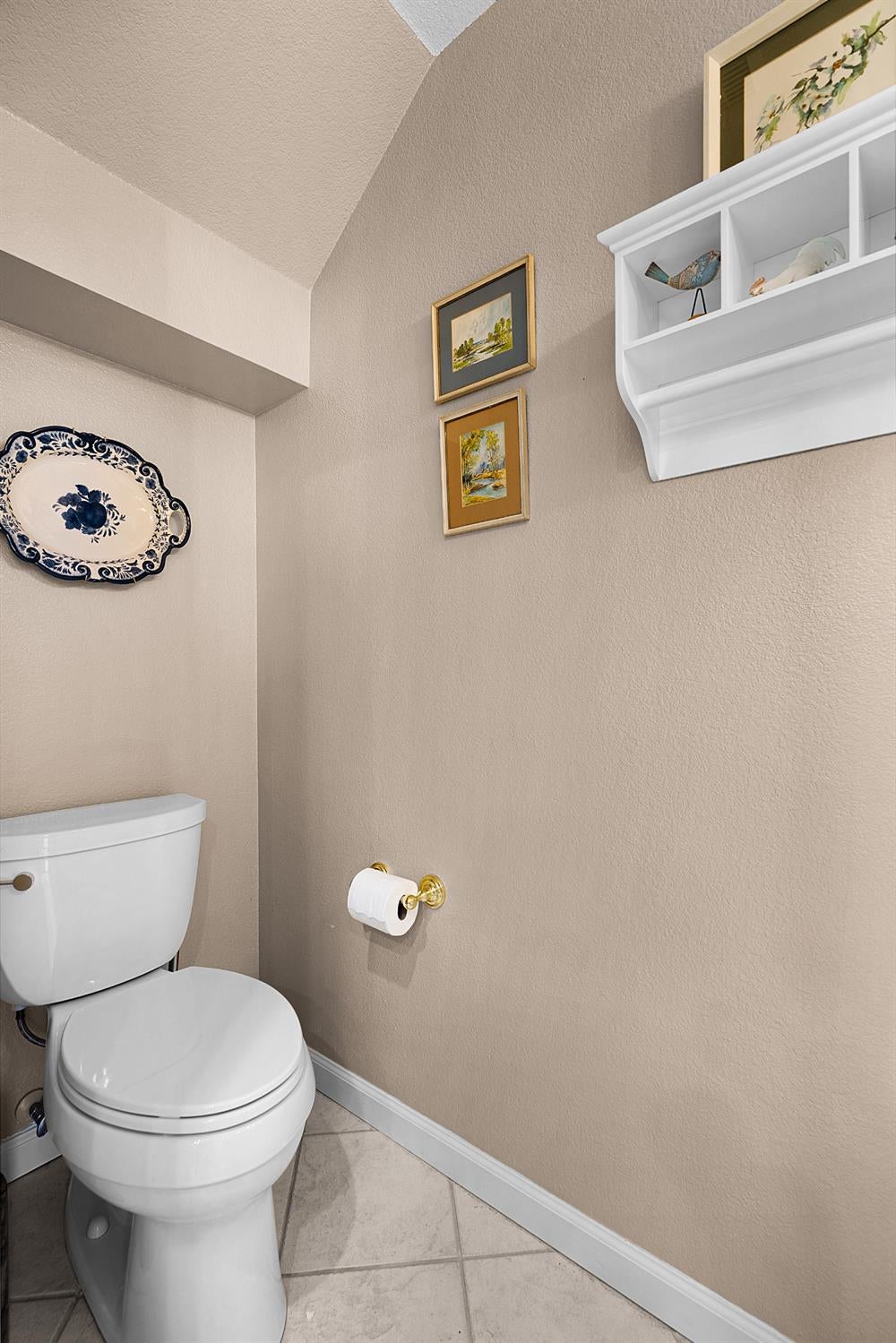 Bathroom with a white toilet, beige walls, framed artwork, and a high white display shelf.