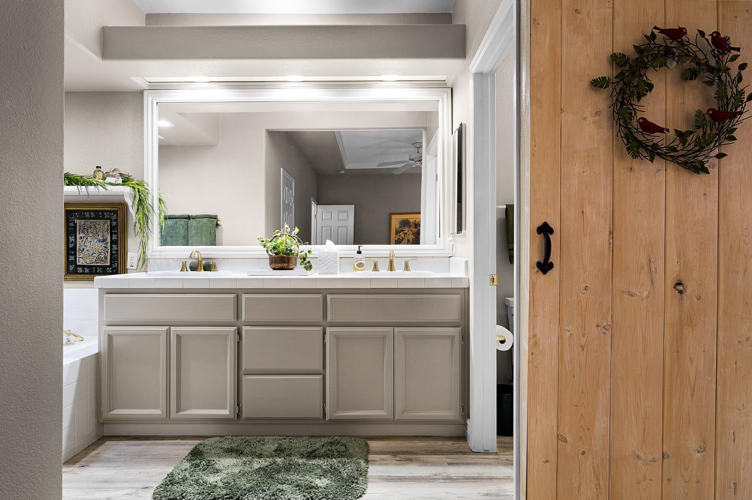 Bathroom vanity with taupe cabinets, brass faucets, a large mirror, and a rustic wood door.
