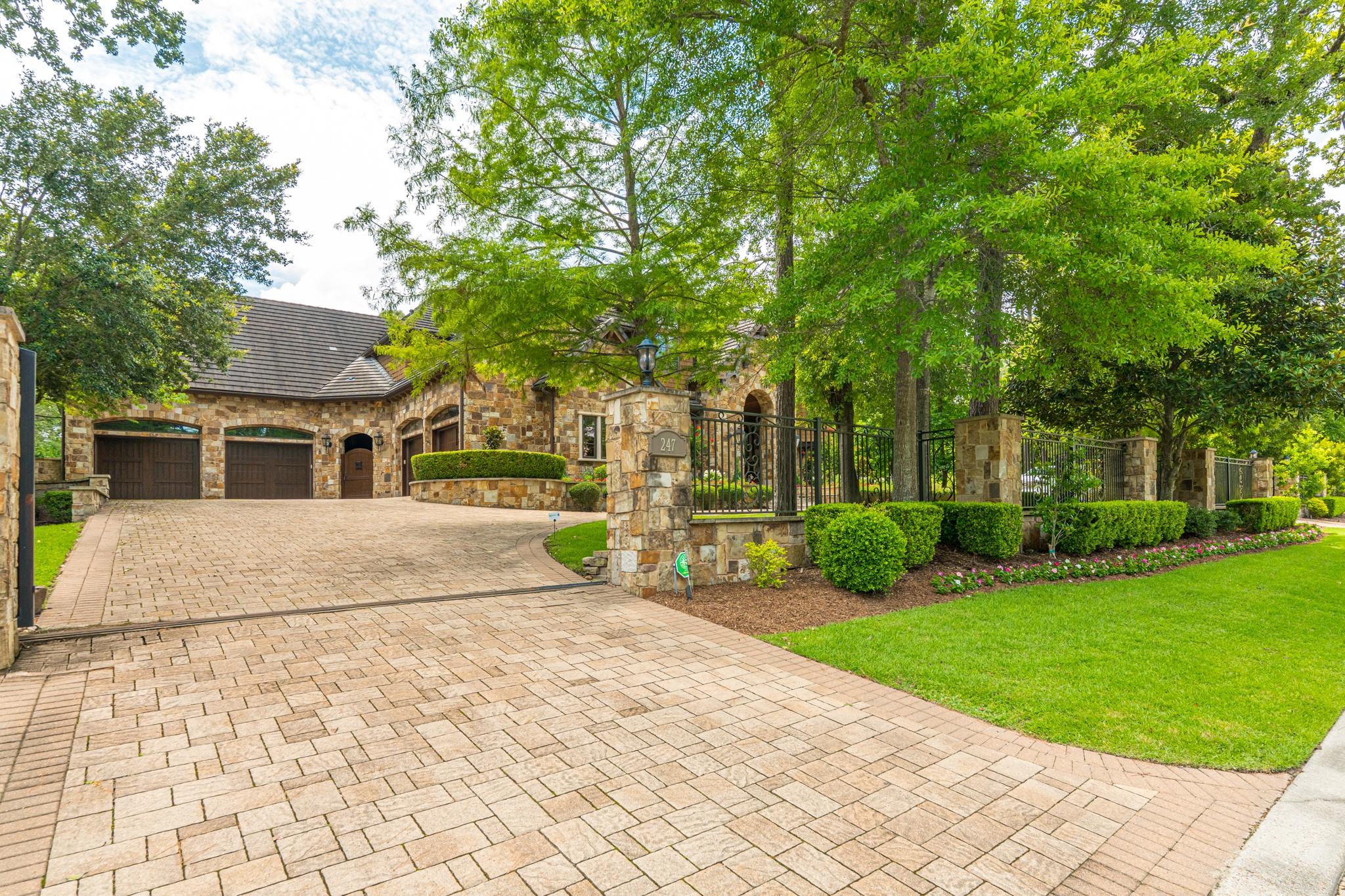 Stone house behind a gated entry, with a broad paver driveway and mature trees.