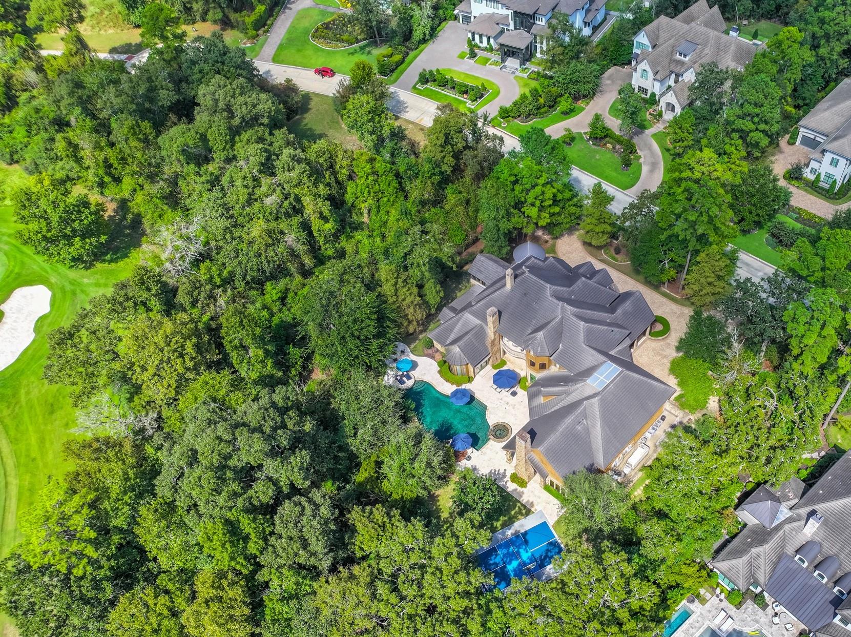 Aerial view of a large home with a swimming pool, tennis court, and wooded grounds.