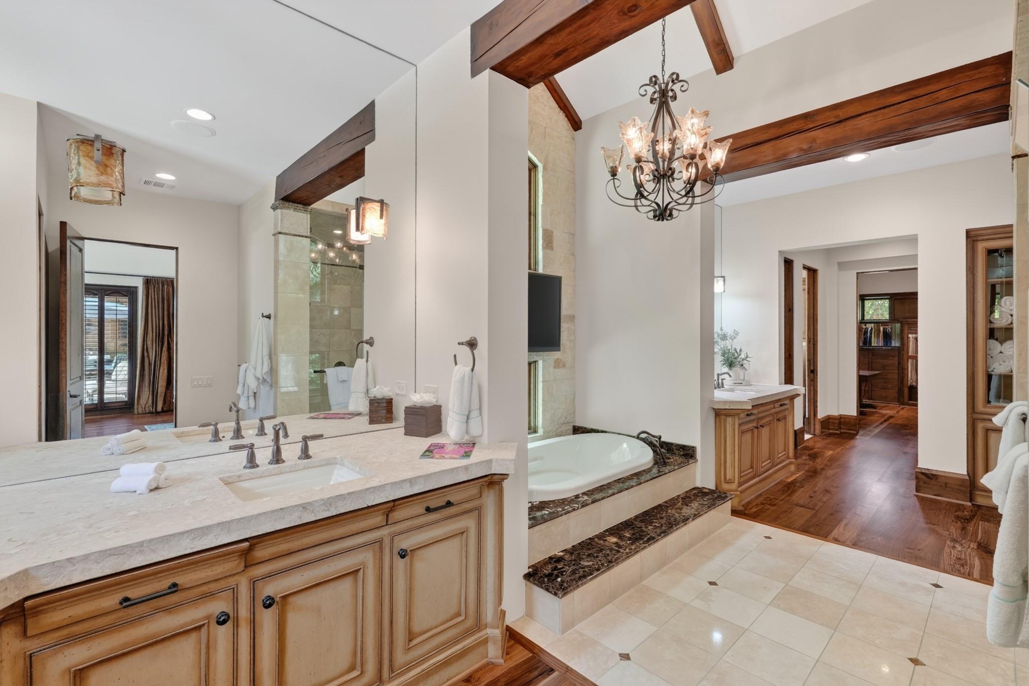 Primary bathroom with a long double vanity, raised soaking tub, exposed wood beams, and chandelier.