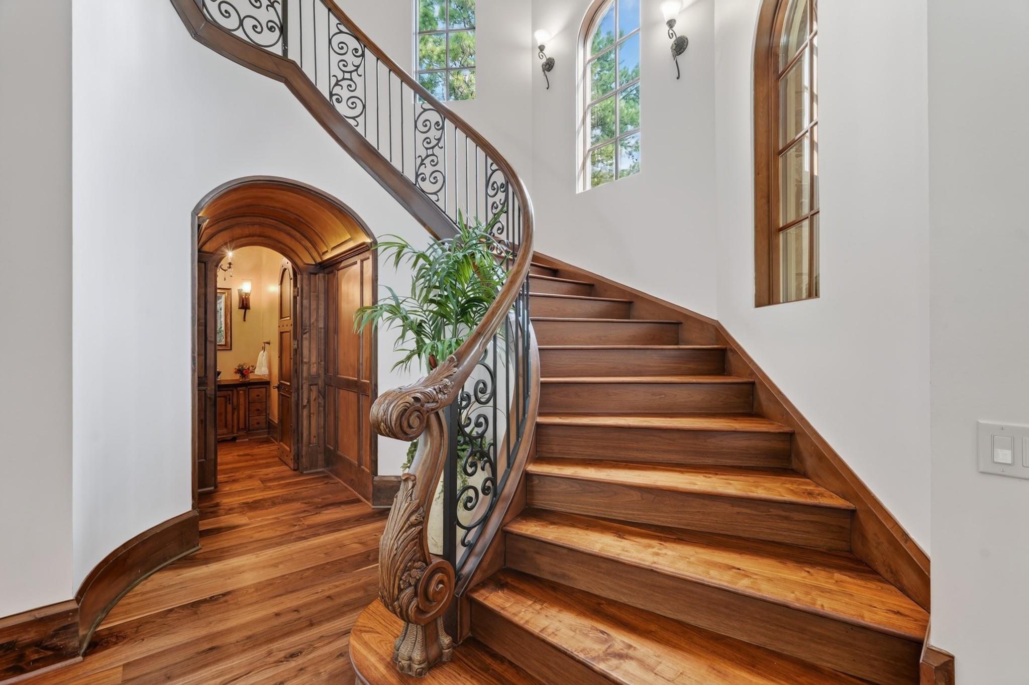 Curved wooden staircase with ornate iron railings, carved newel posts, and tall arched windows.