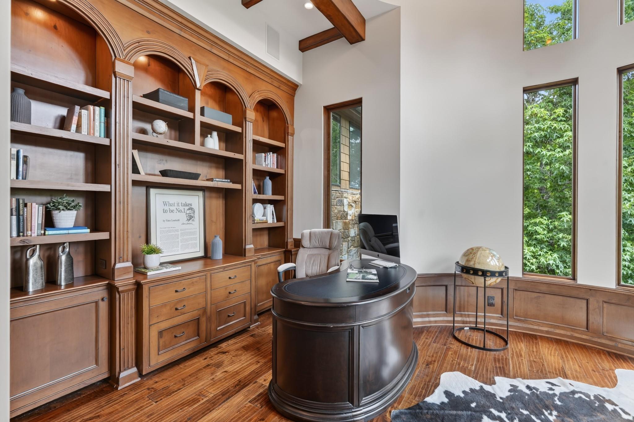 Home office with arched wood bookcases, a curved desk, exposed beams, and tall windows.