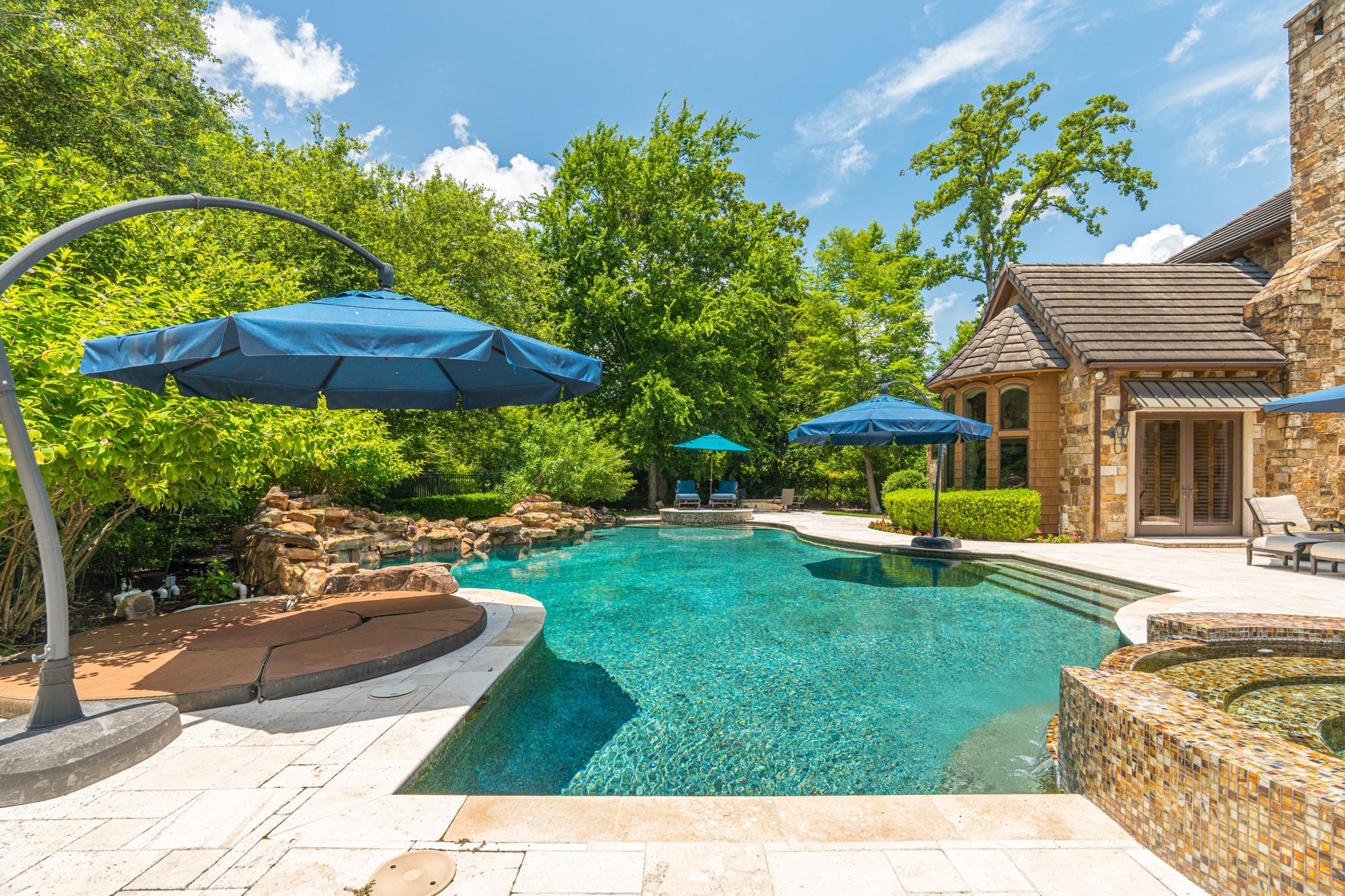 Backyard swimming pool beside a stone house, with a tiled spa, rock waterfall, and blue umbrellas.