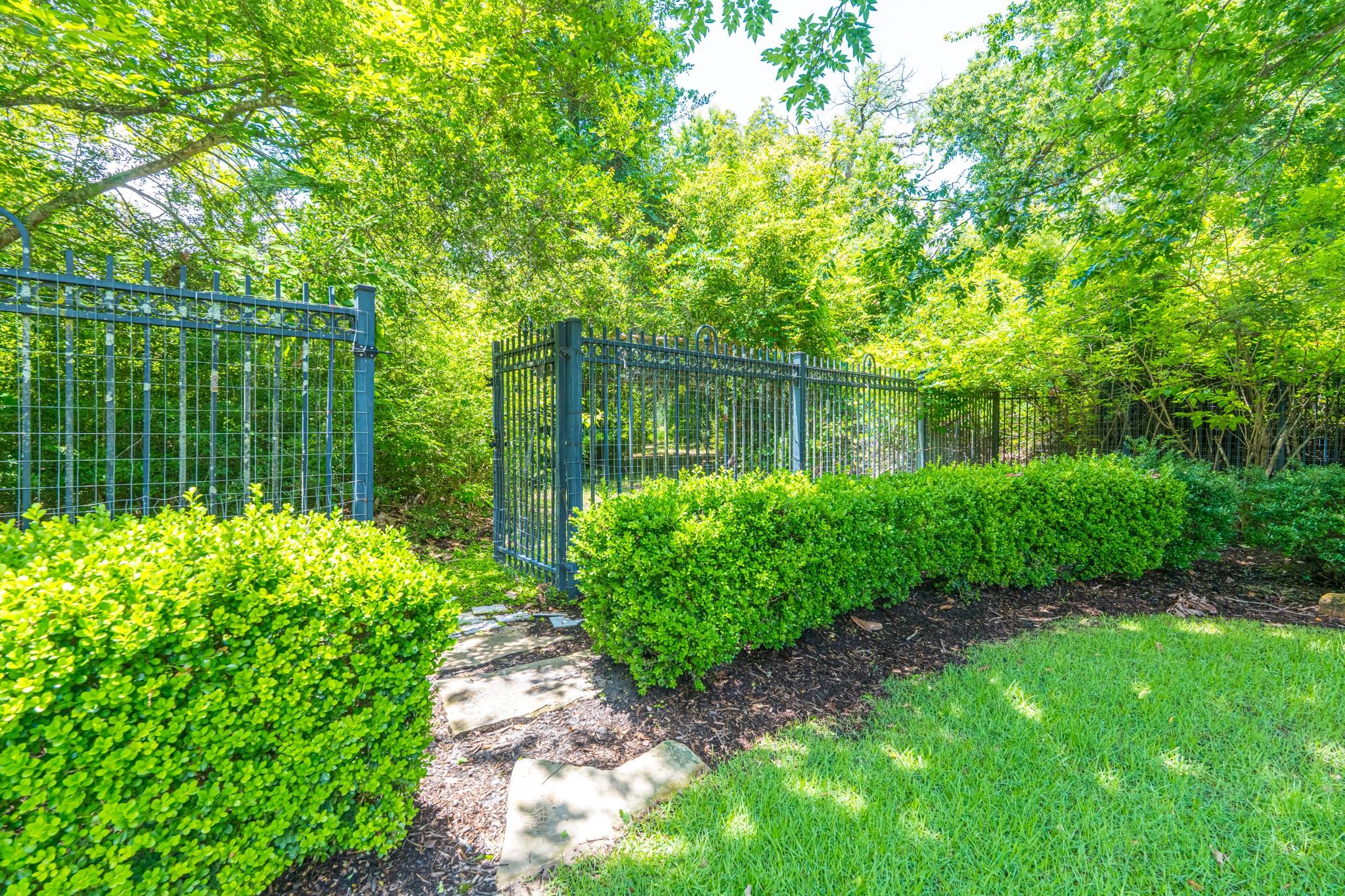Landscaped yard with a black metal gate, clipped hedges, stone path, and mature trees.
