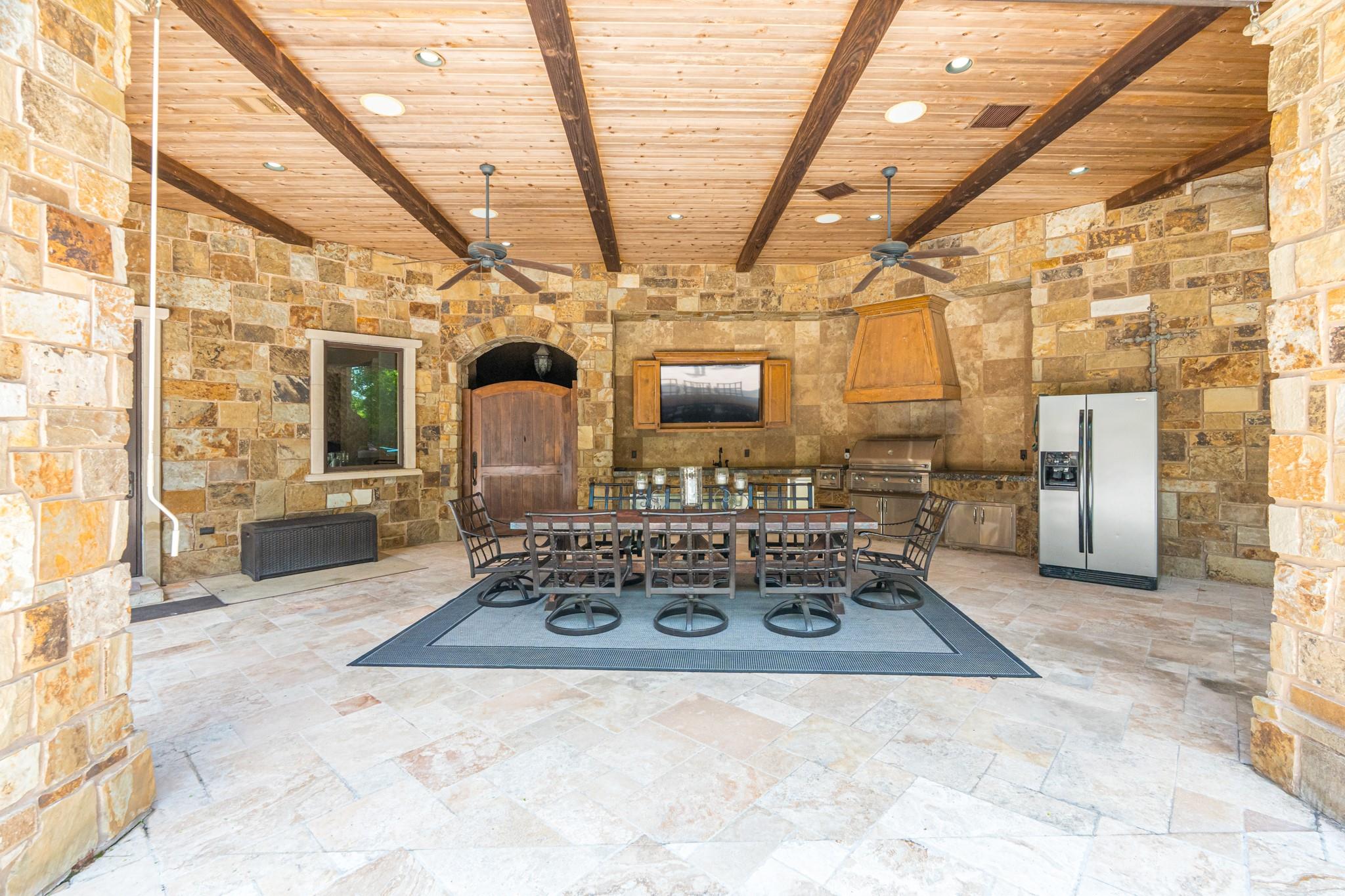 Covered outdoor kitchen with a stone dining area, built-in grill, and exposed wood ceiling beams.