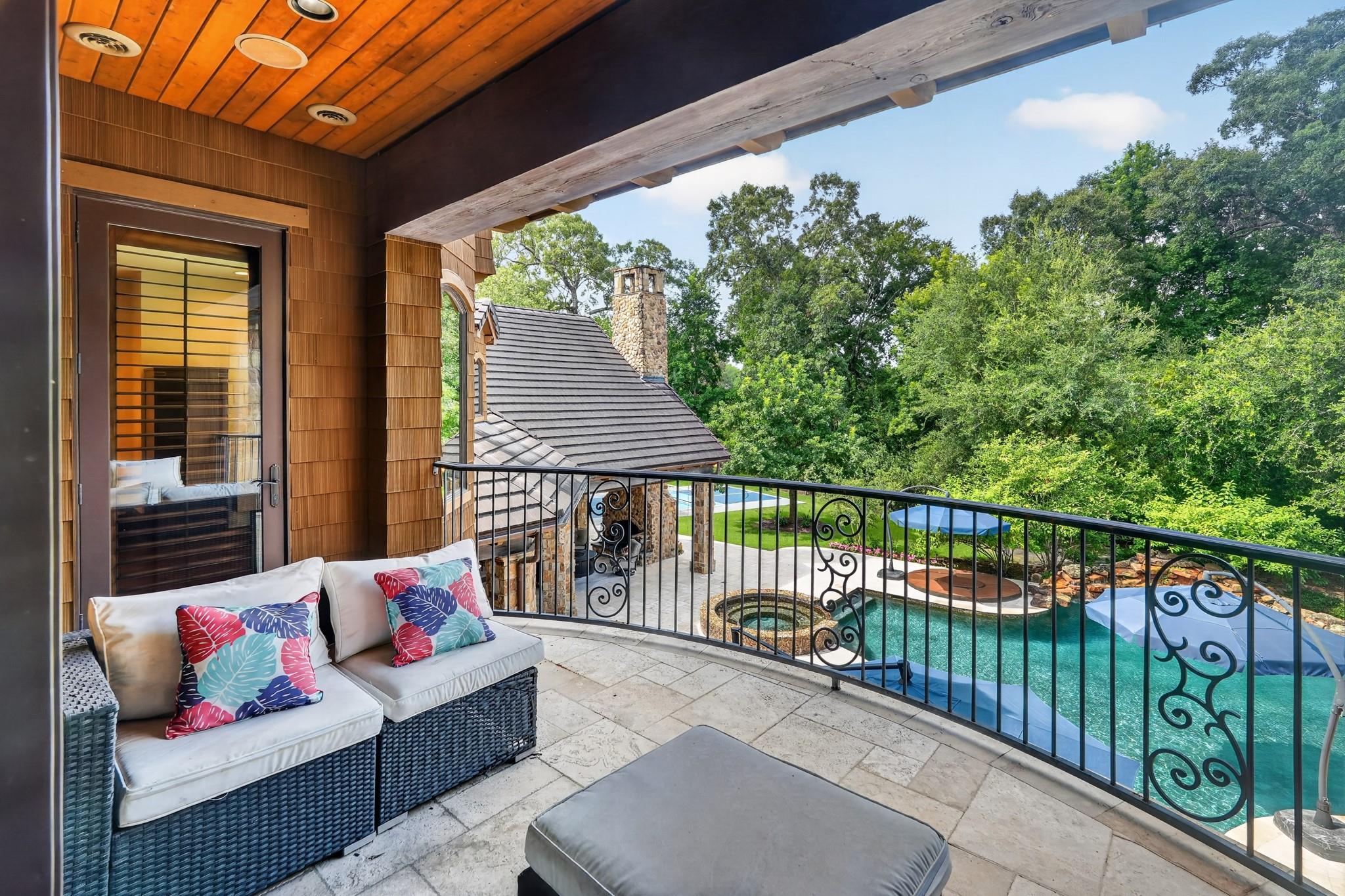 Covered balcony with wicker seating and colorful cushions overlooking a pool and wooded grounds.
