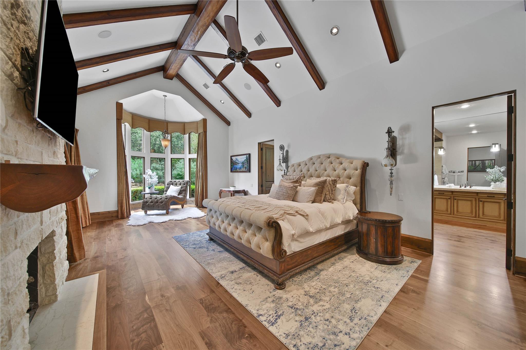Furnished bedroom with a tufted bed, vaulted wood-beam ceiling, stone fireplace, and tall bay windows.