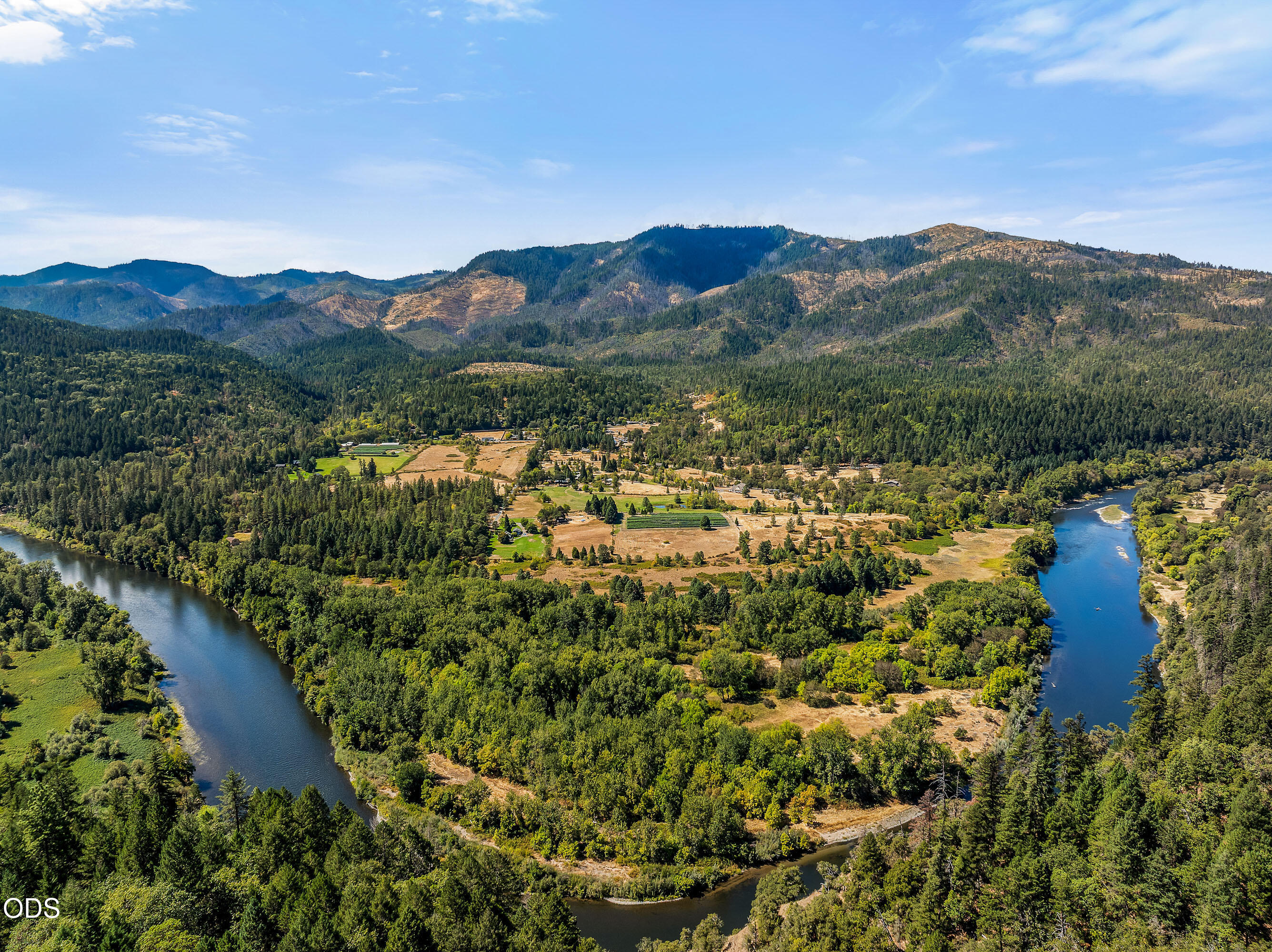 Aerial view of a forested river valley with open fields and mountains beyond.