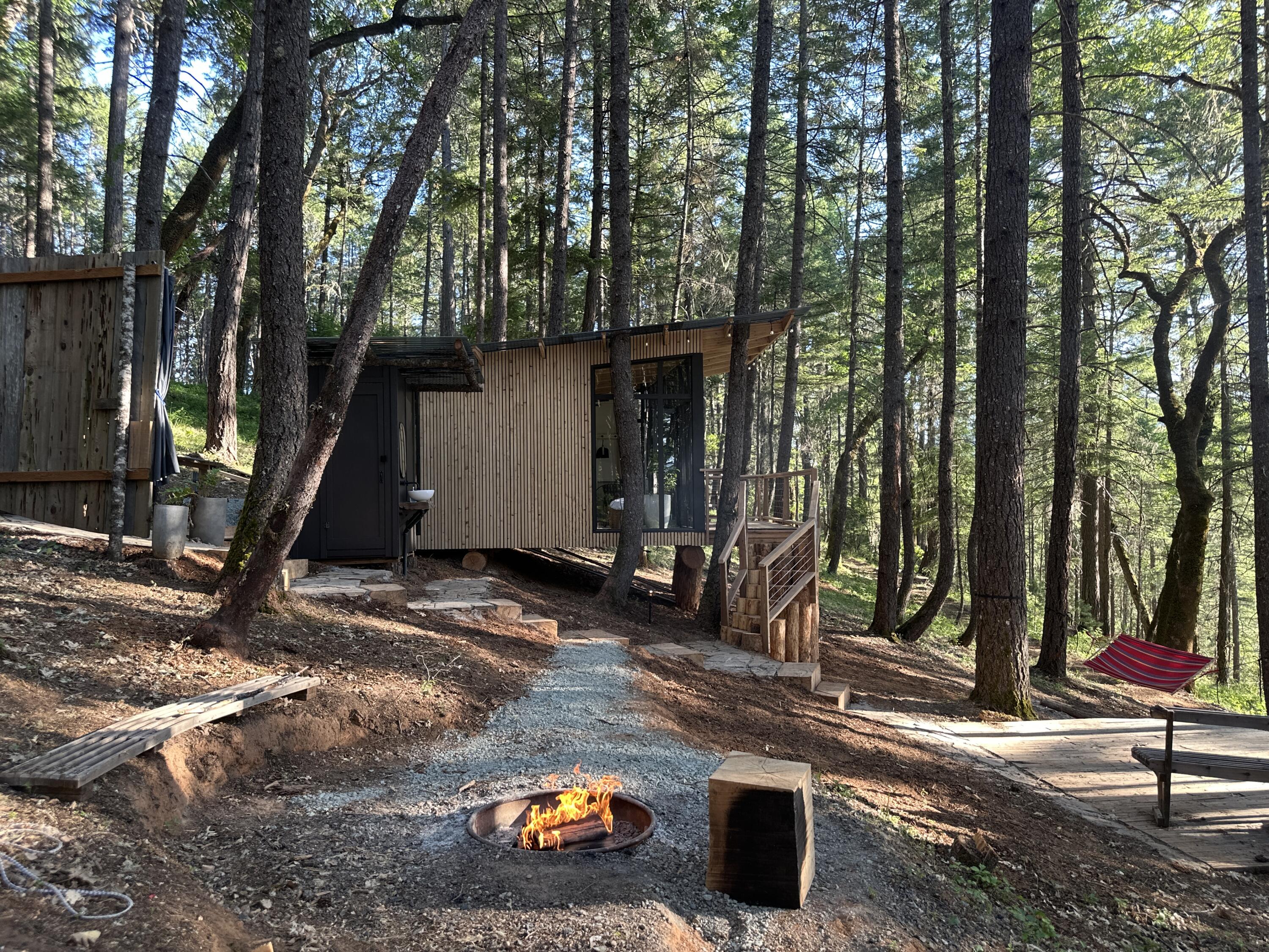 Modern wood-sided cabin in a forest, with a deck, fire pit, and red hammock.