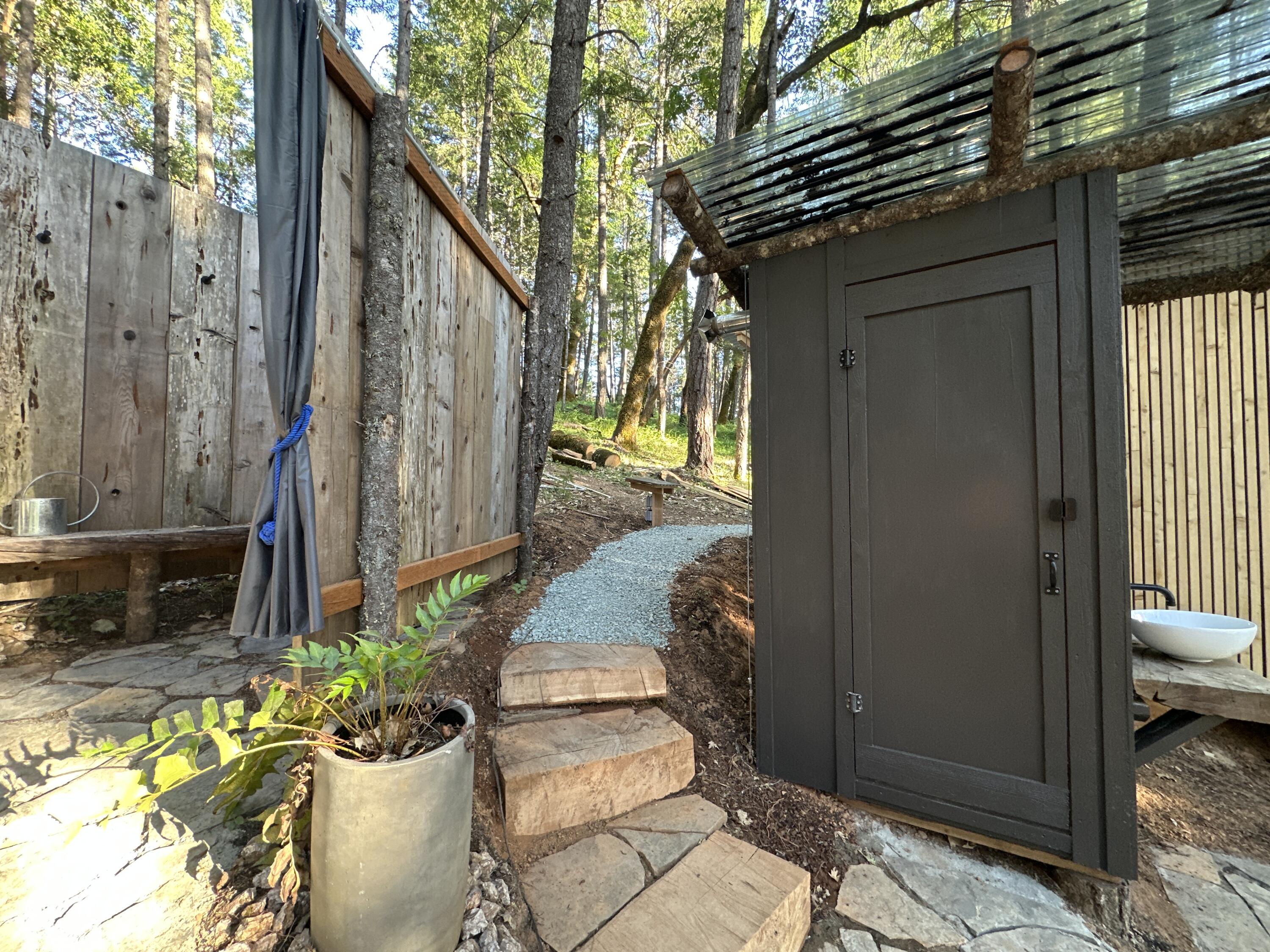 Wooded outdoor bathroom with a dark door, white basin, and stone steps.