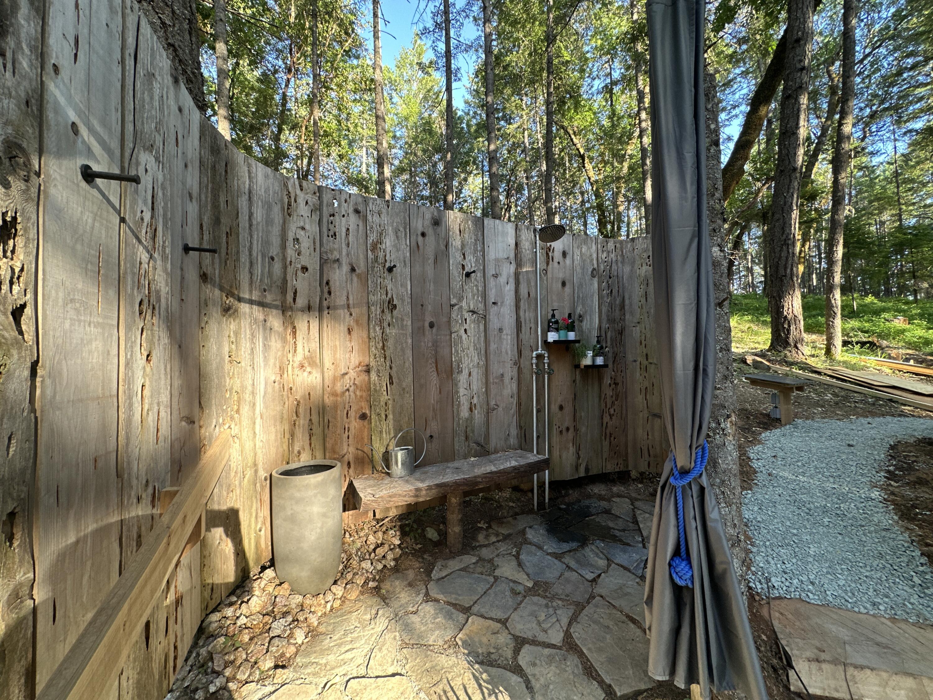 Rustic outdoor shower beside a forest, with a plank privacy fence, stone paving, and a wooden bench.