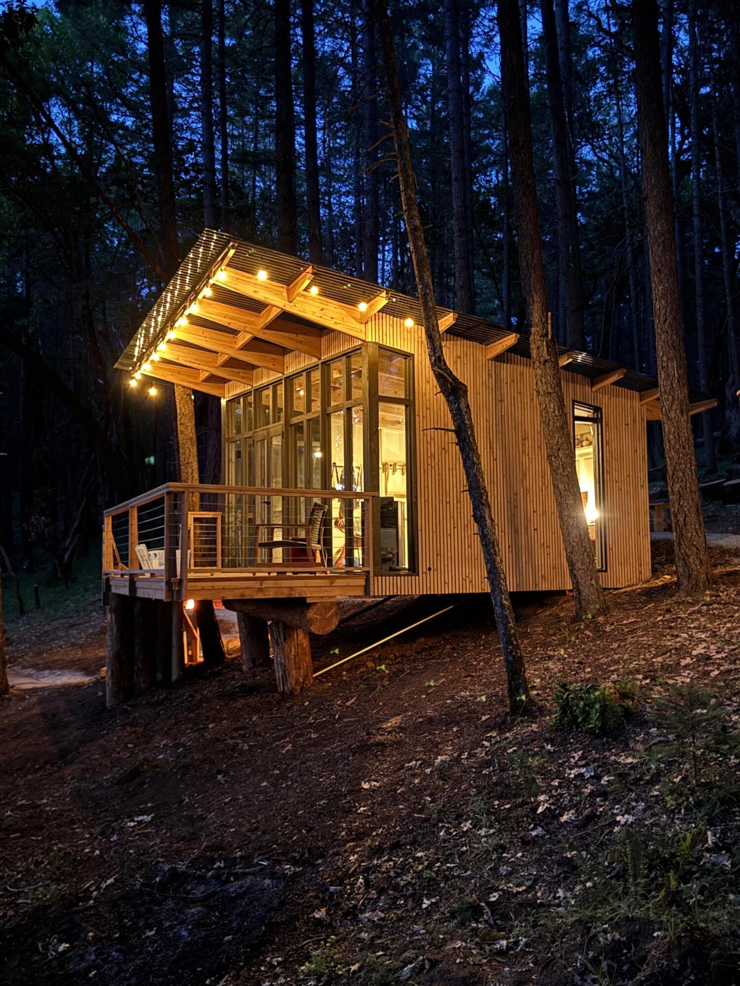 Elevated timber cabin glowing among trees, with a broad roof and glass-fronted deck.