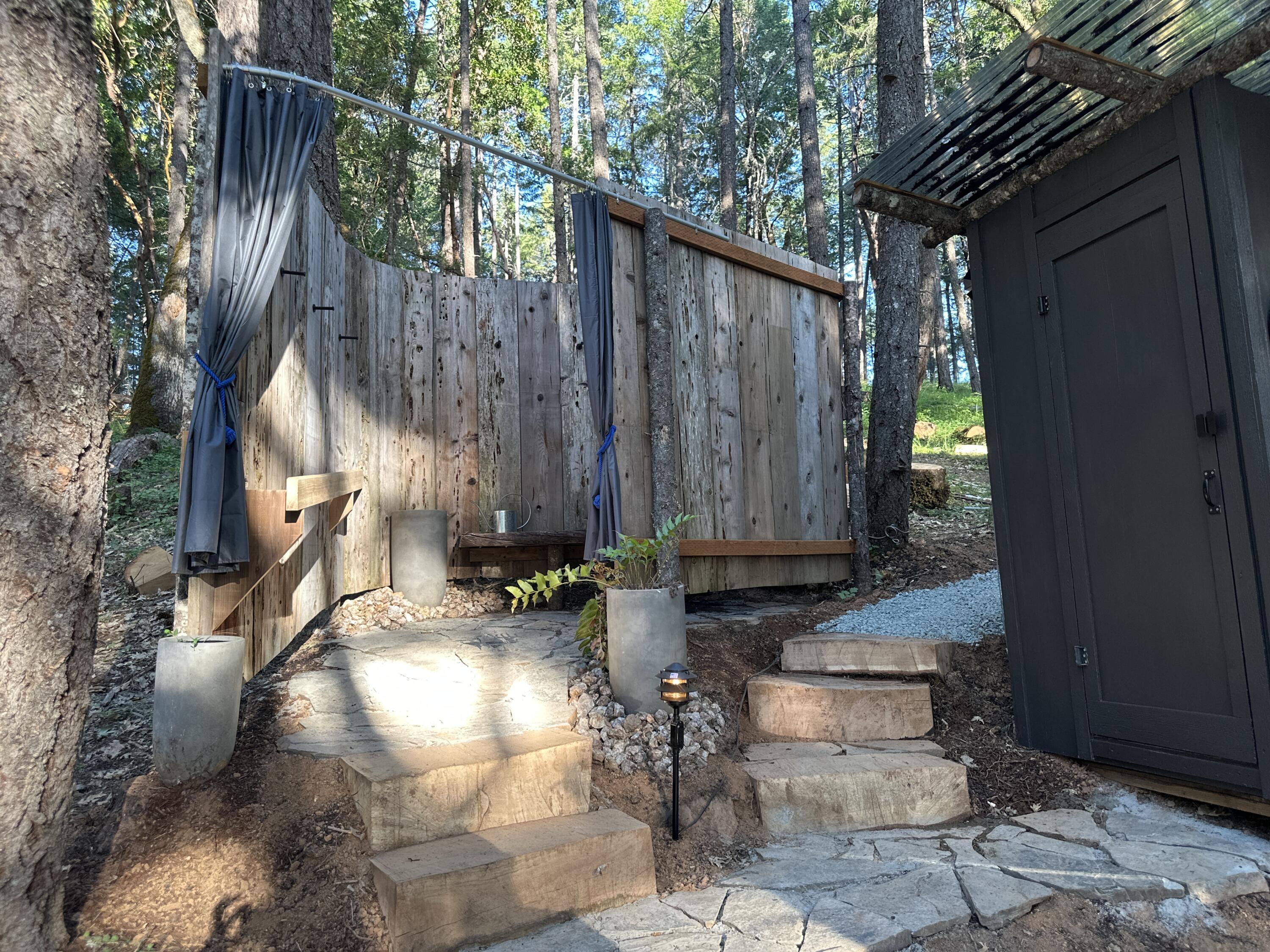 Outdoor shower with wooden privacy screens and gray curtains beside a dark shed in a wooded setting.