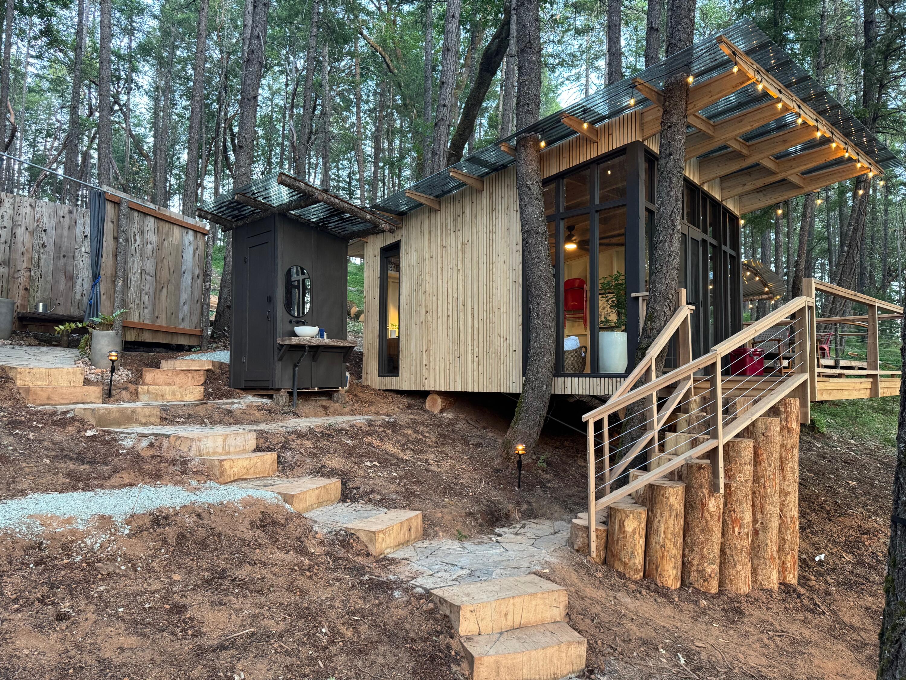 Modern wood-sided cabin among tall trees, with a raised deck, string lights, and stone steps.