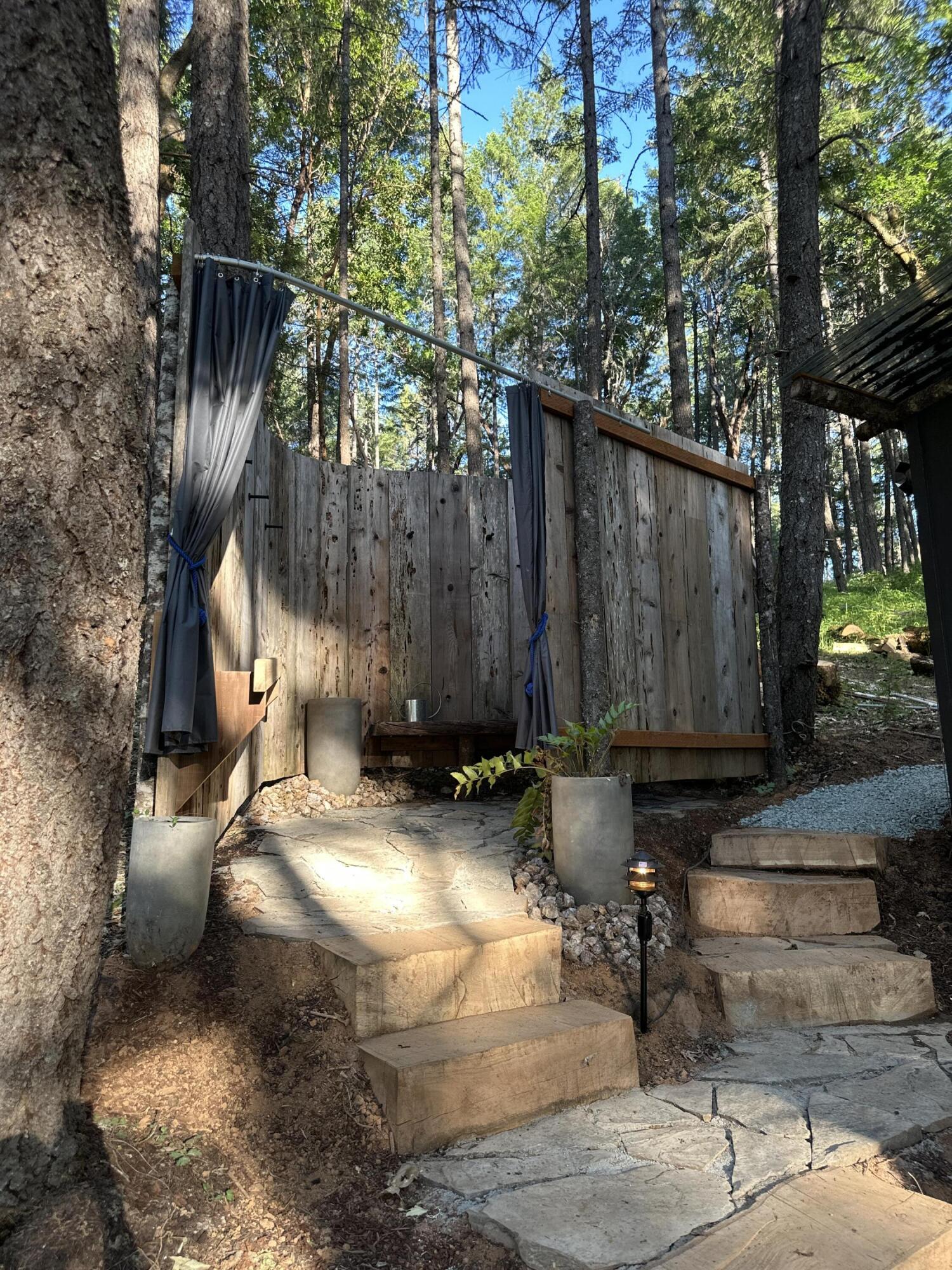 Rustic outdoor shower among trees, with wooden walls, dark curtains, and stone steps.