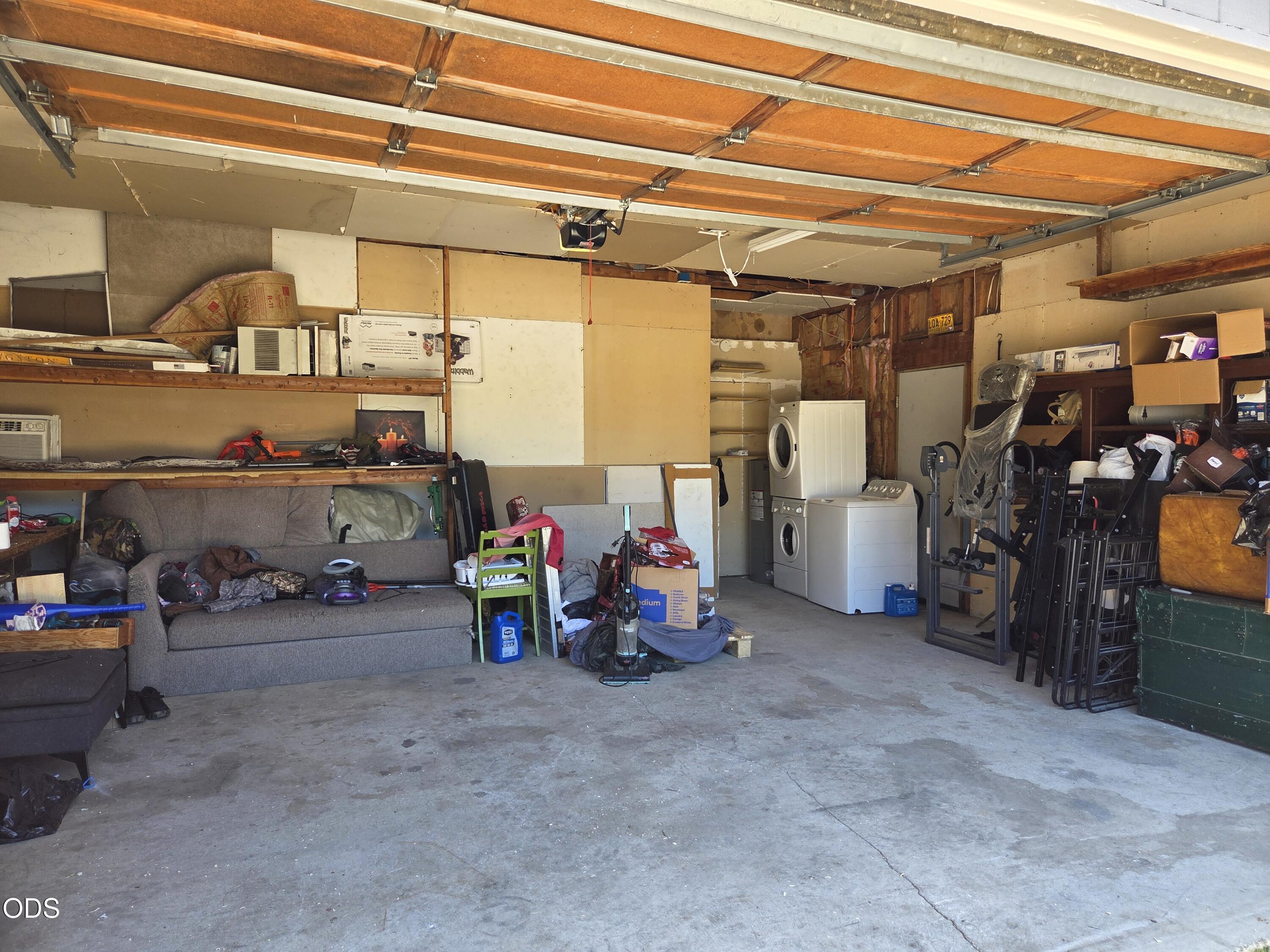 Cluttered garage with an open wooden door, storage shelves, a sofa, and stacked washer and dryer.