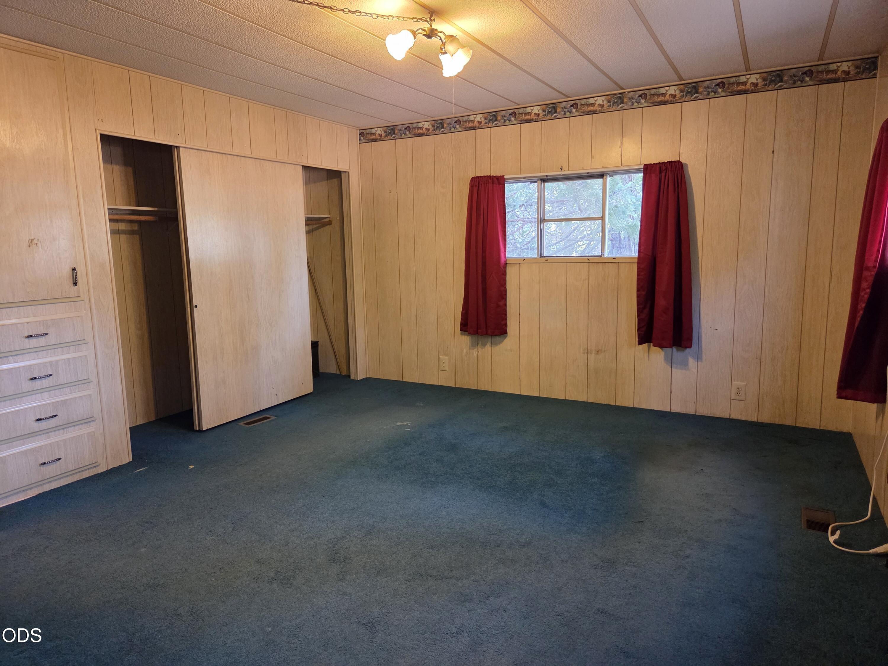 Empty bedroom with wood paneling, blue carpet, built-in closet, and red curtains.