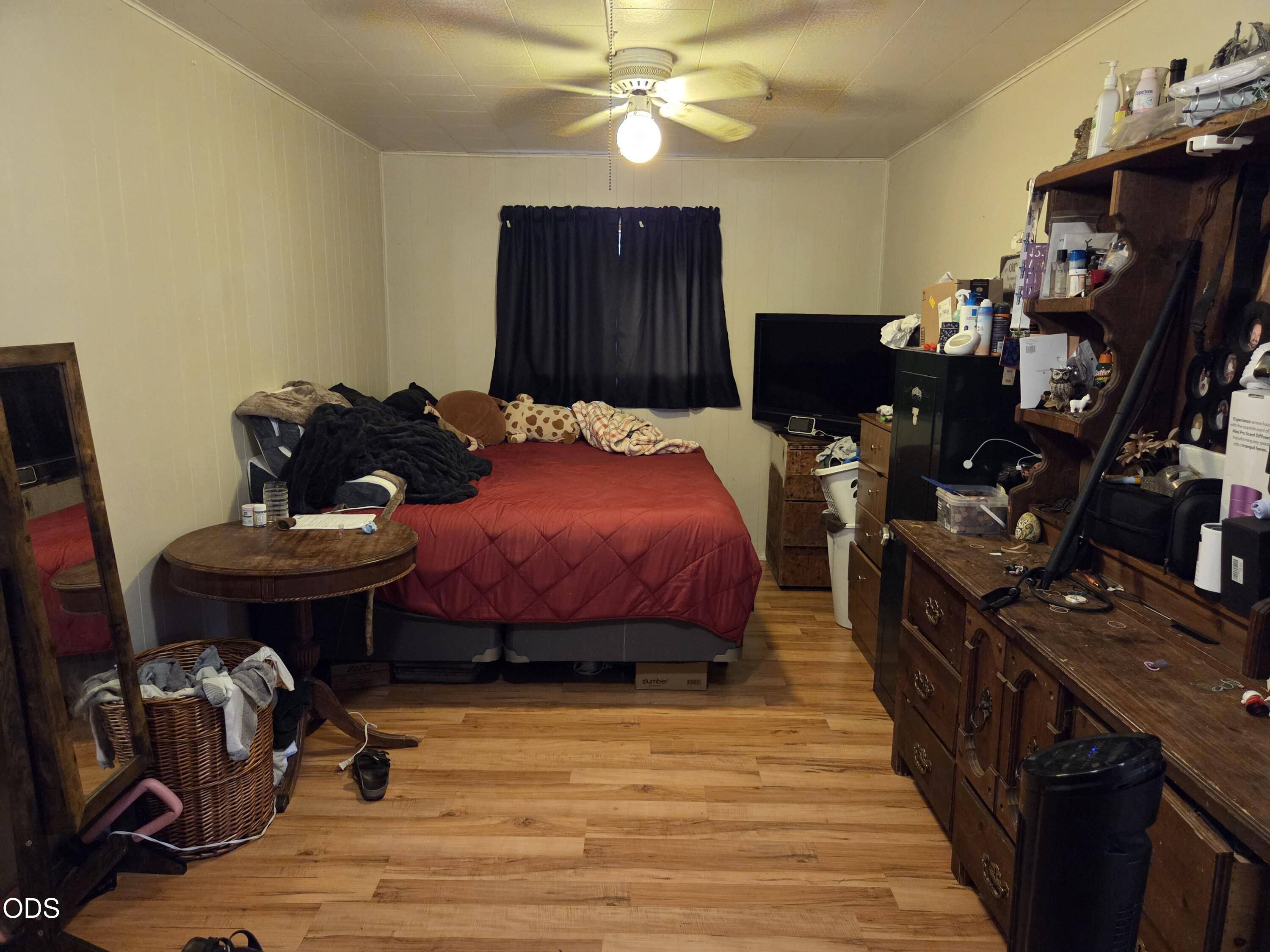 Bedroom with a red quilted bed, dark curtains, and crowded wooden dressers.
