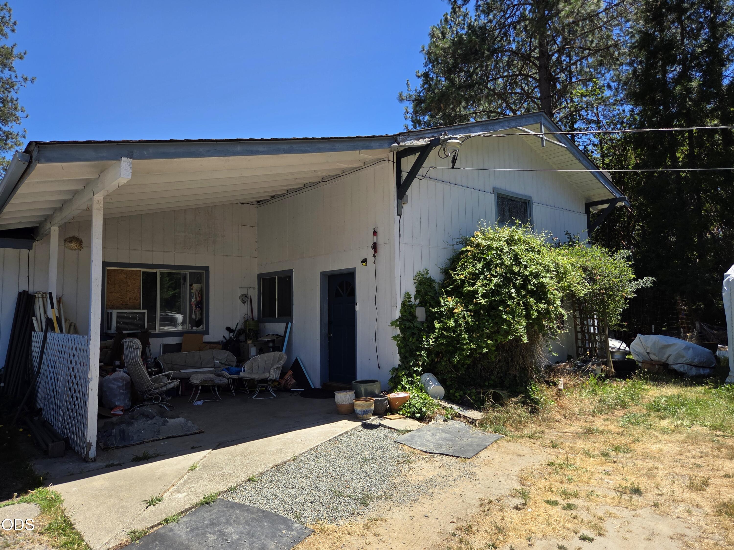 White house with a deep covered carport, blue door, and cluttered patio.