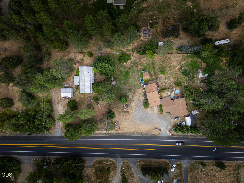 Aerial view of a rural home and outbuildings beside a two-lane road, surrounded by trees.