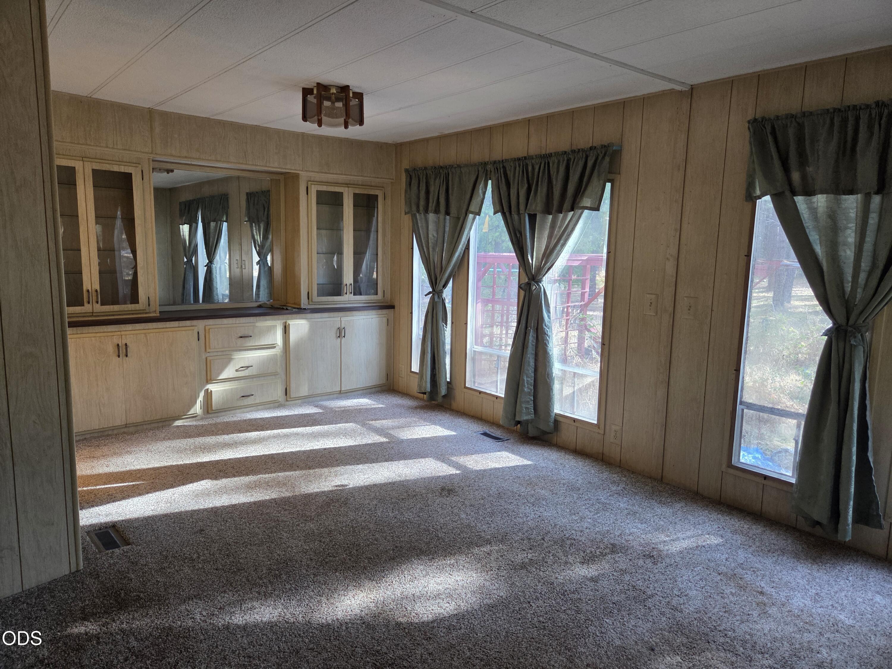 Empty carpeted room with built-in wood cabinets and tall curtained windows.