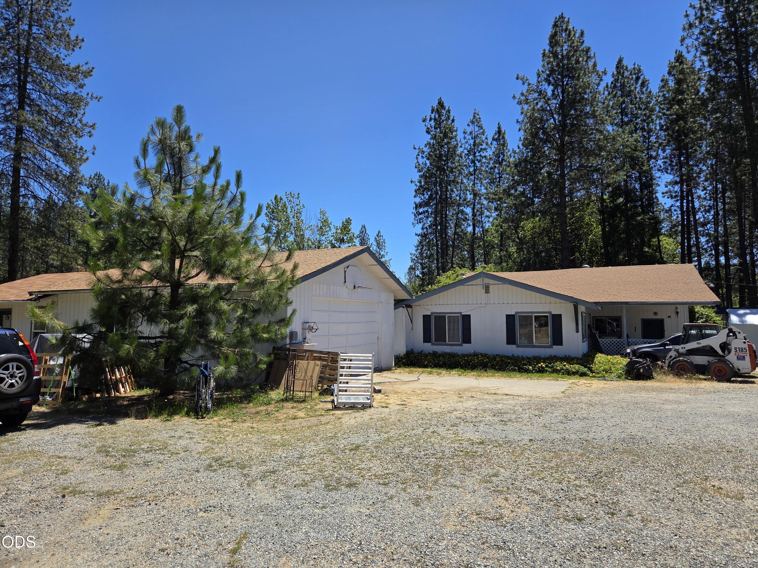 White single-story home with garage and gravel driveway among tall pine trees.