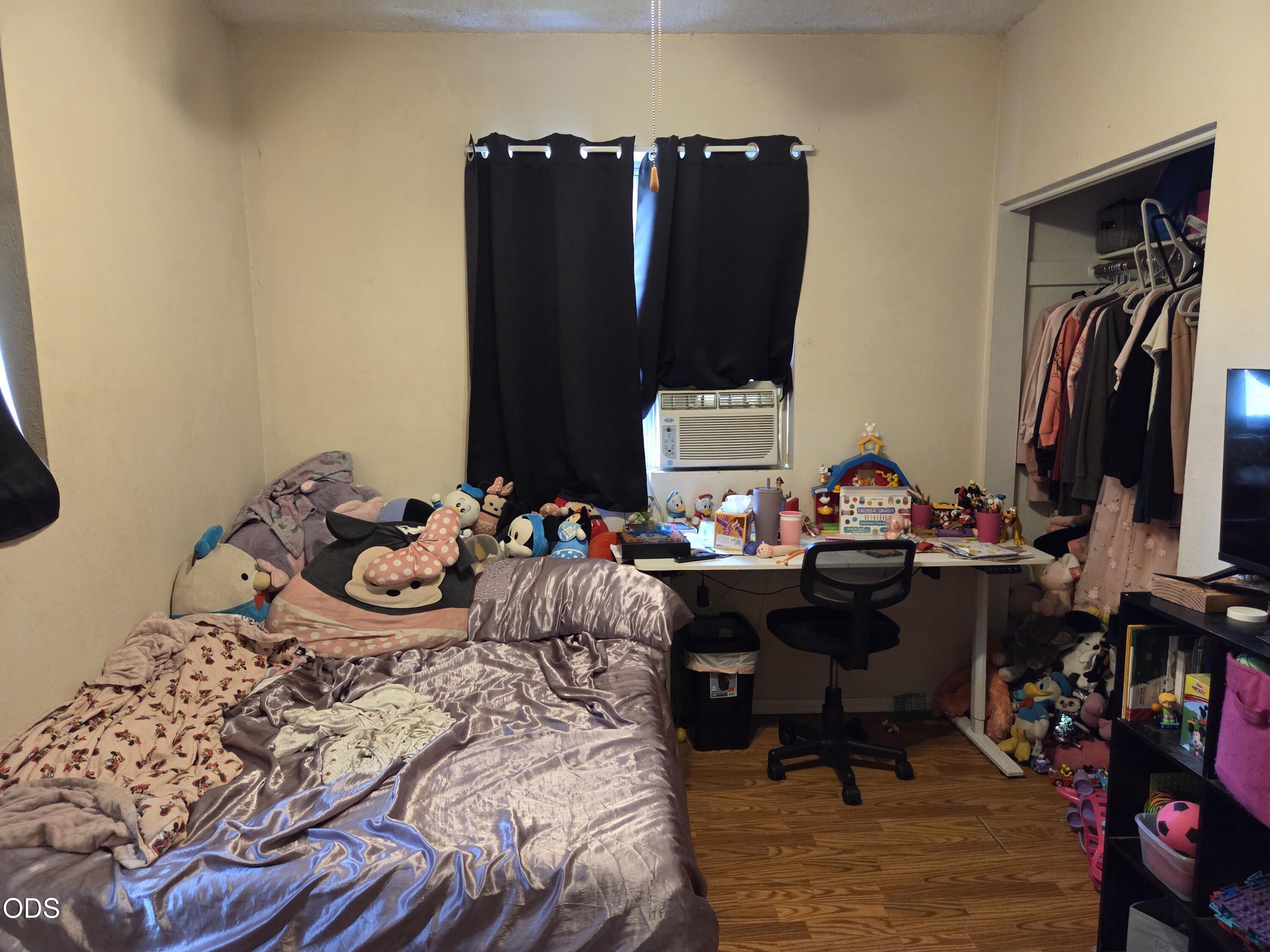 Bedroom with an unmade satin-covered bed, stuffed toys, a crowded desk, and an open clothes closet.
