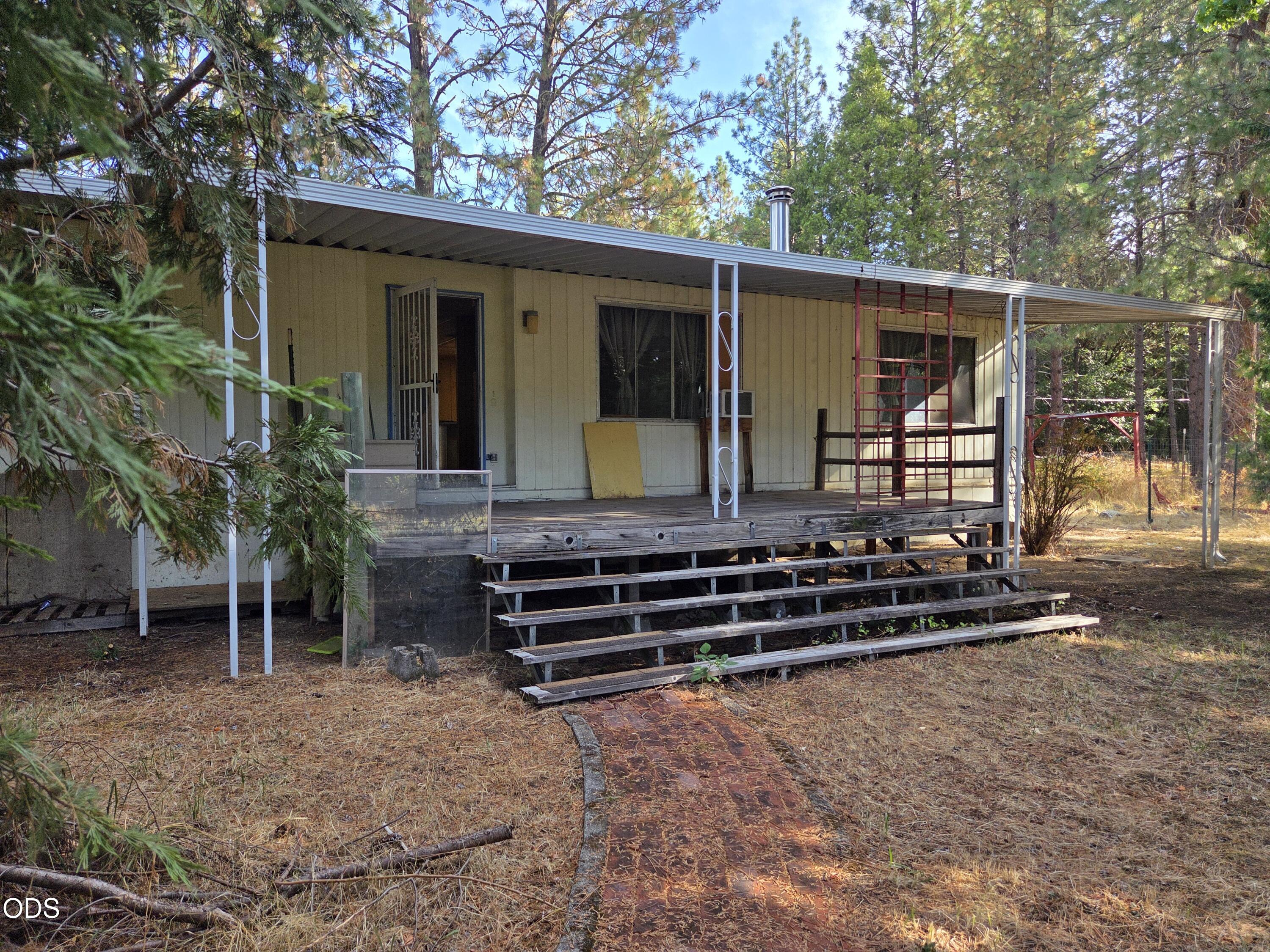 Single-story house with a long covered porch, weathered steps, and red scaffolding among pine trees.