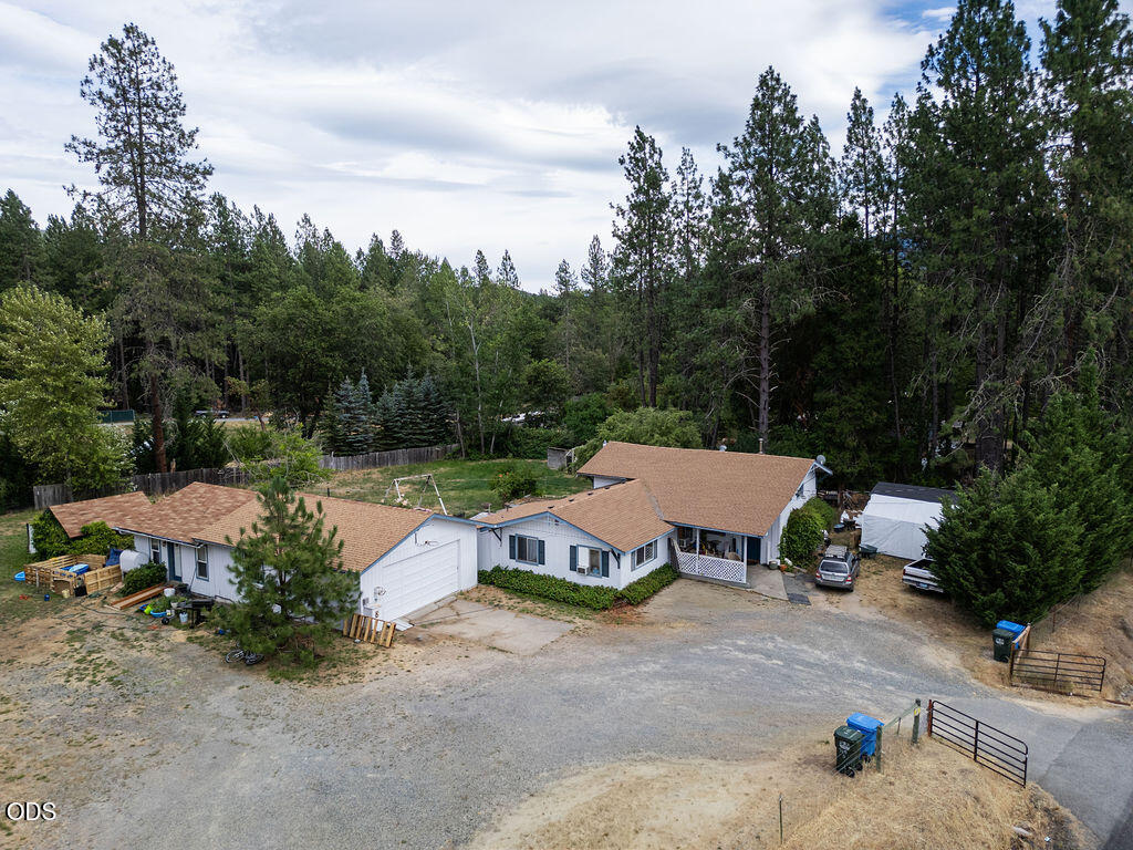 Aerial view of a white home with tan roofs, gravel drive, and surrounding pine trees.
