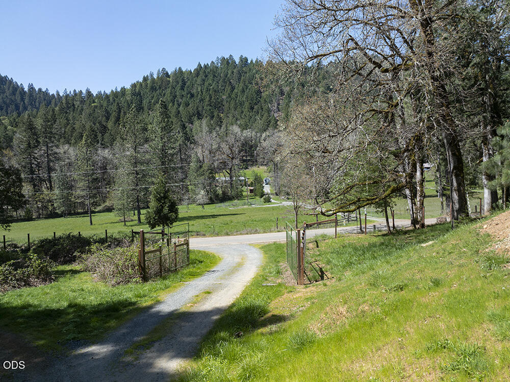 Gravel driveway through an open gate, overlooking green pasture and forested hills.