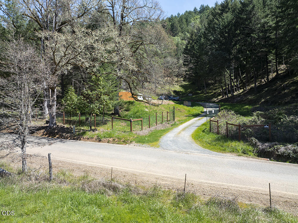 Rural driveway entrance with fencing, wooded hillside, and a small orange outbuilding.