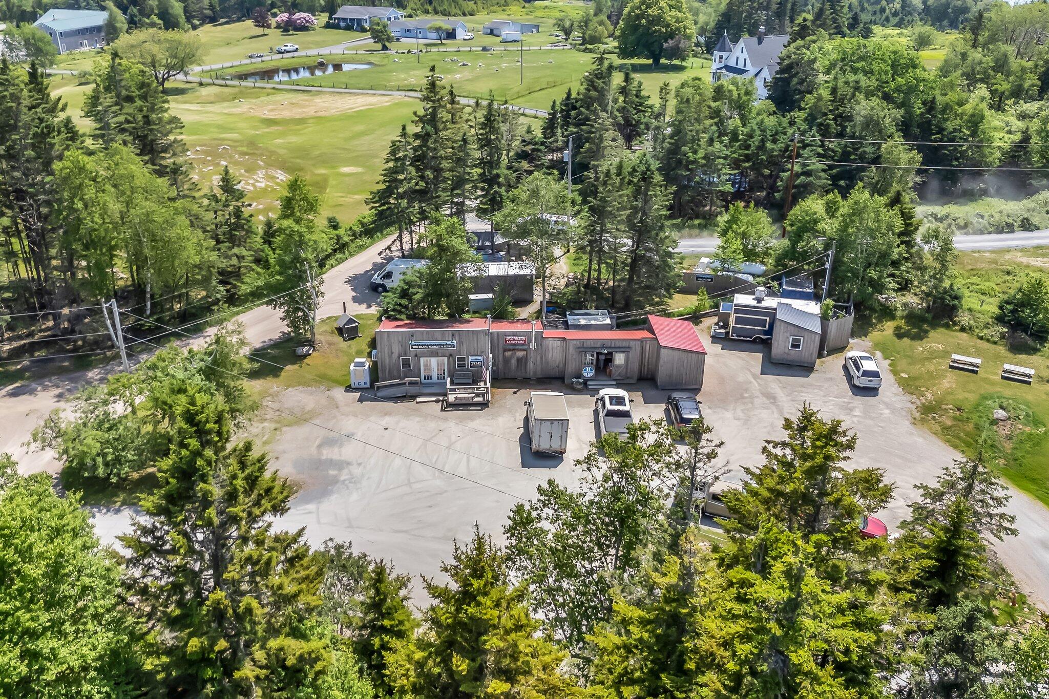 Aerial view of a roadside commercial market with red roofs, parking area, and wooded surroundings.