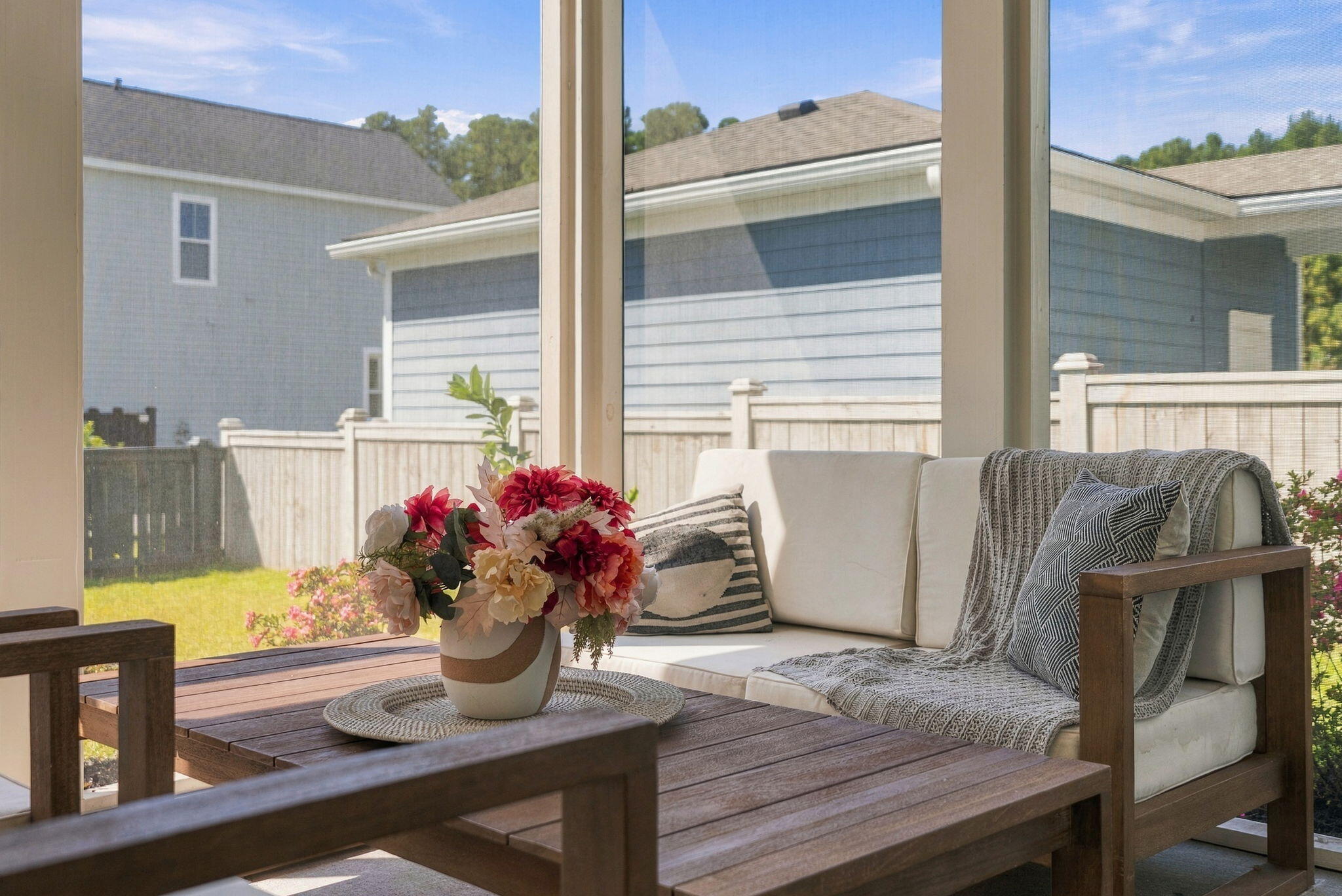 Furnished sunroom with a wooden sofa and flower arrangement beside large windows overlooking a fenced yard.