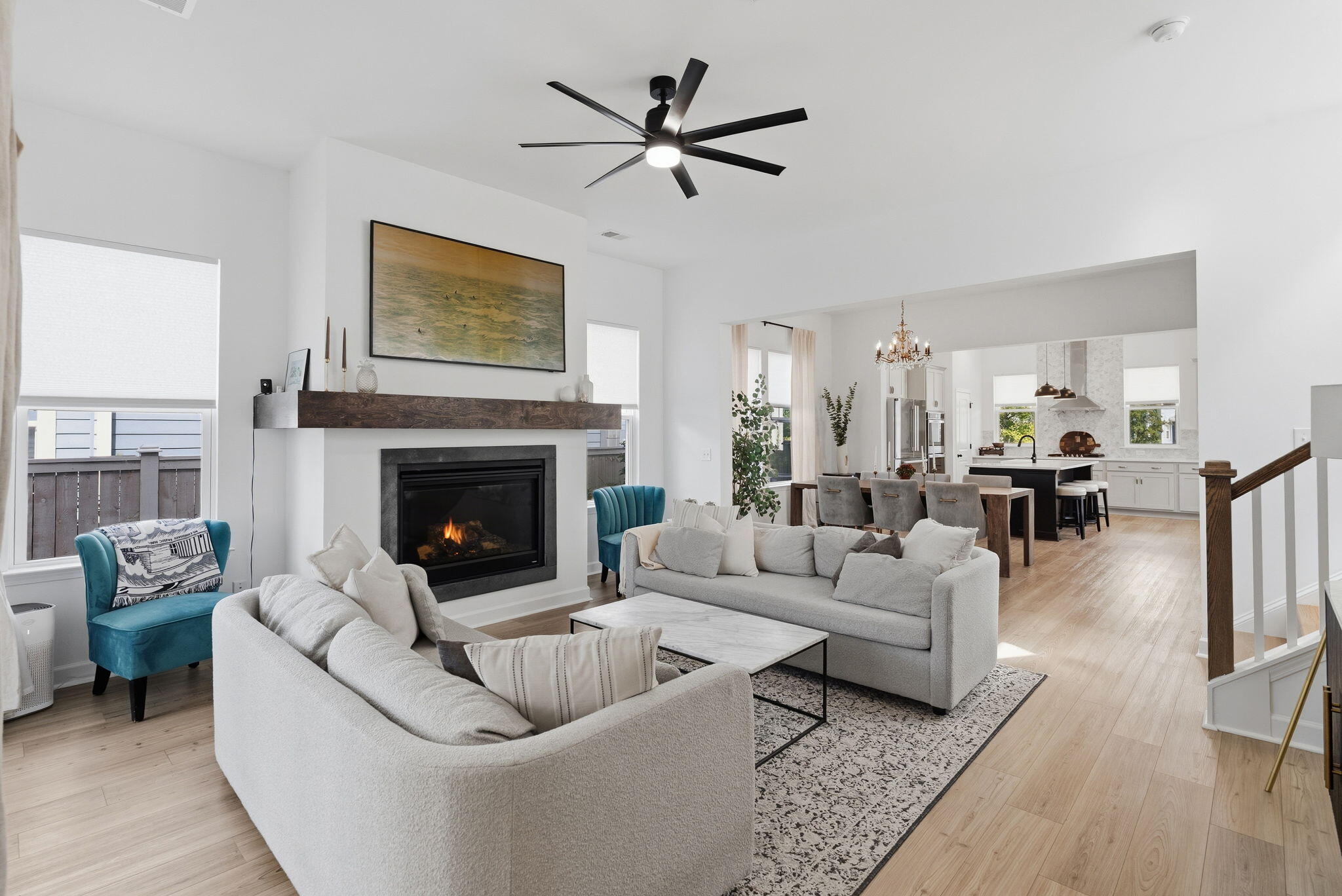 Open-plan living room with a lit fireplace, pale sofas, teal chair, and adjoining kitchen.