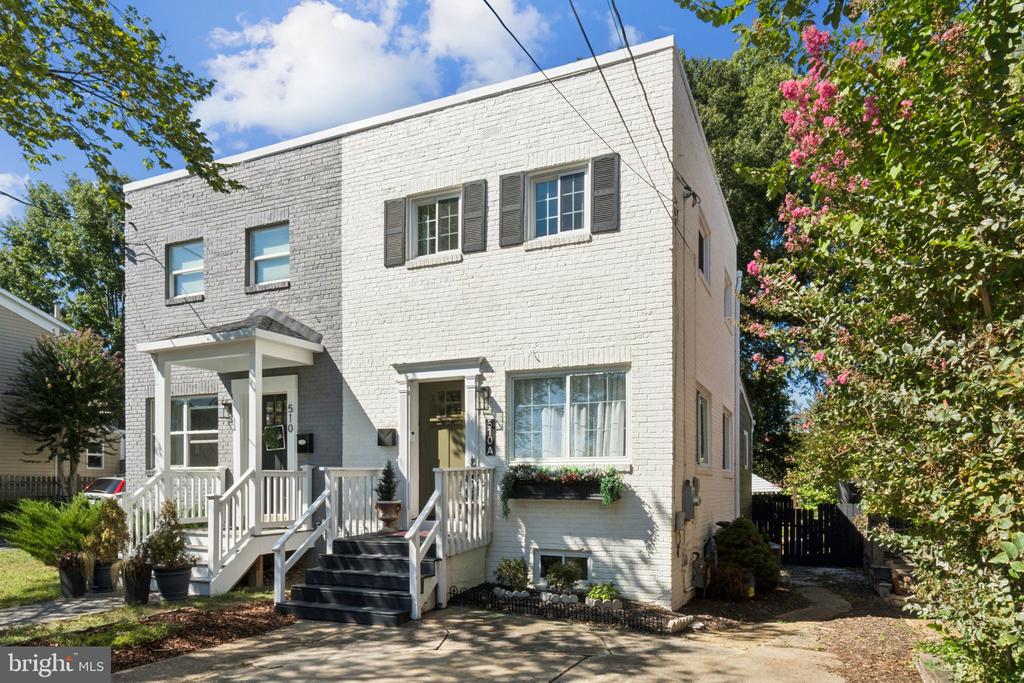 White brick two-story house with covered front porches, white railings, and mature trees.
