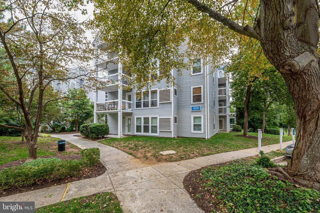 Gray condominium building with balconies, framed by mature trees and a curving sidewalk.