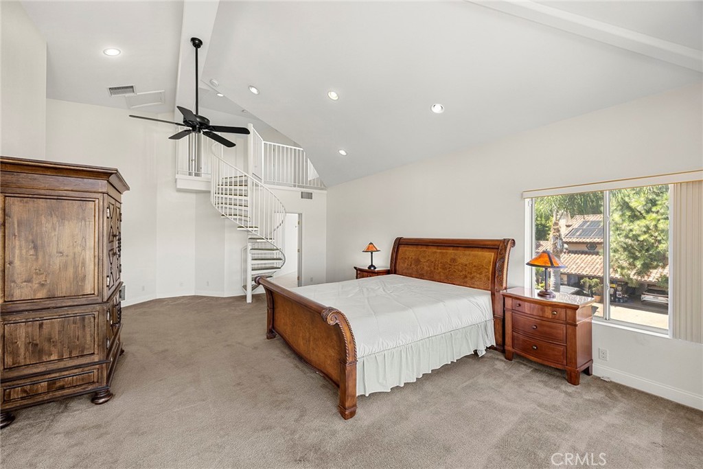 Furnished bedroom with a white spiral staircase, vaulted ceiling, and large window overlooking trees.