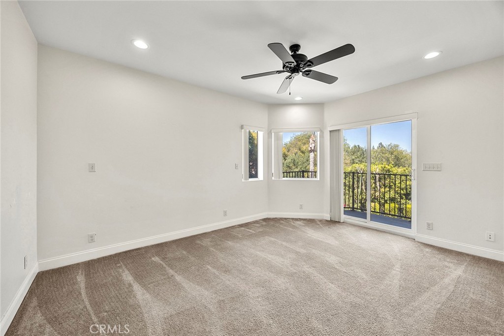 Empty carpeted bedroom with a ceiling fan and sliding glass door to a balcony overlooking trees.