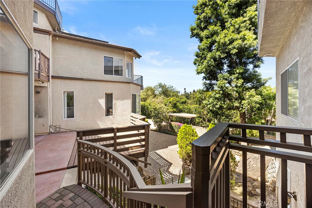 Courtyard patio between beige residences, with wooden railings, paved steps, shrubs, and a large leafy tree.