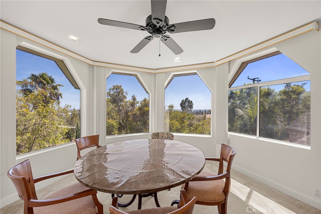 Dining nook with an oval wooden table, ceiling fan, and angled bay windows overlooking trees.