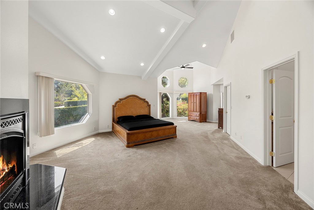 Spacious bedroom with vaulted ceilings, a wooden bed, large windows, and a lit fireplace.