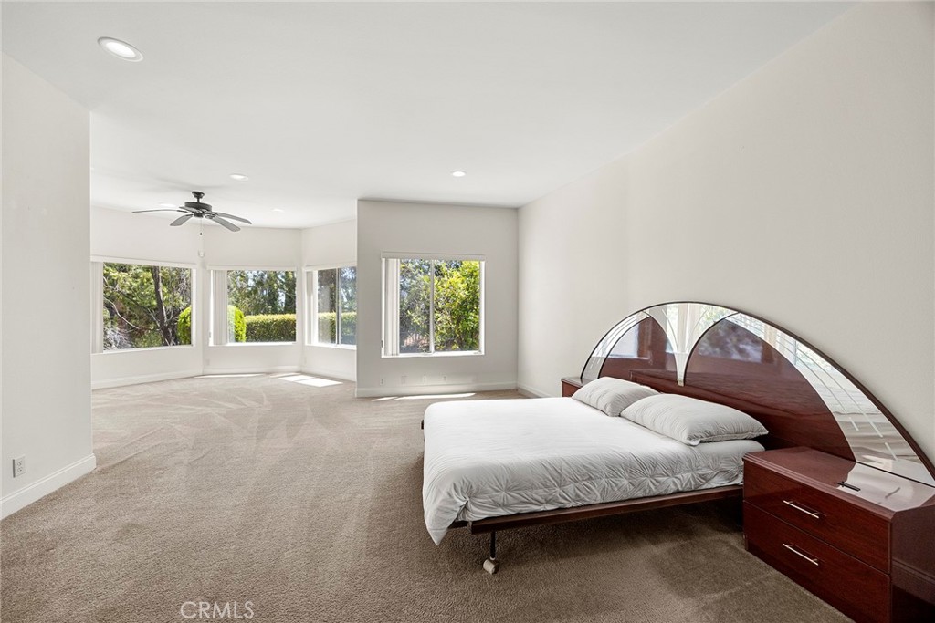 Spacious carpeted bedroom with a white bed, curved wood headboard, and large windows overlooking greenery.
