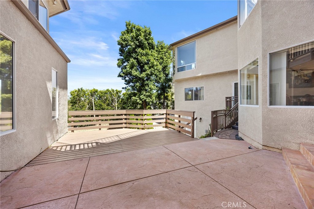 Concrete patio and raised wooden deck between stucco walls, with mature trees beyond.