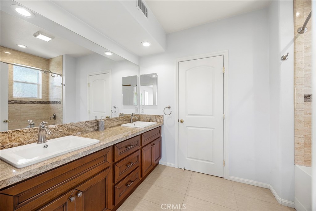 Bathroom with a double-sink granite vanity, dark wood cabinetry, and a tiled shower beside a window.