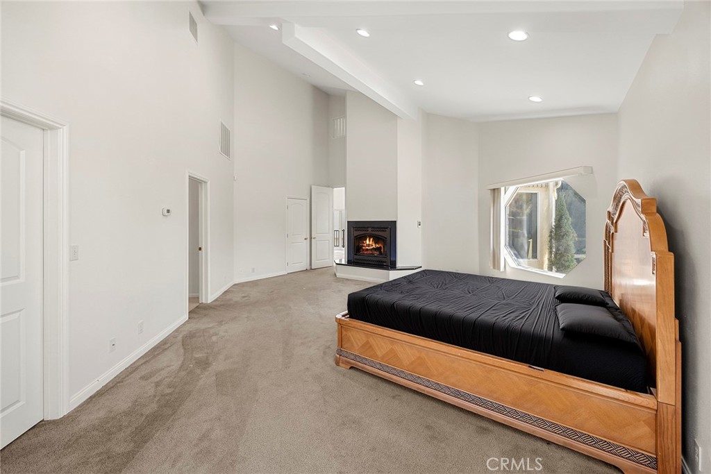 Spacious bedroom with a wooden bed, beige carpet, tall ceiling, and a fireplace.