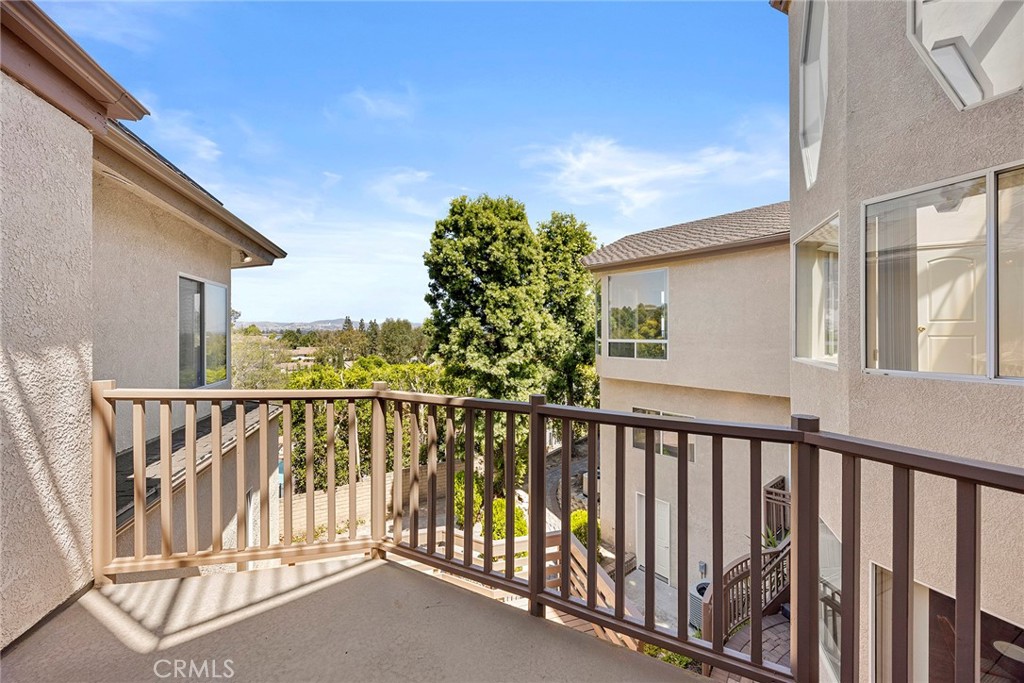 Second-story balcony with brown railing overlooking leafy trees and neighboring homes.