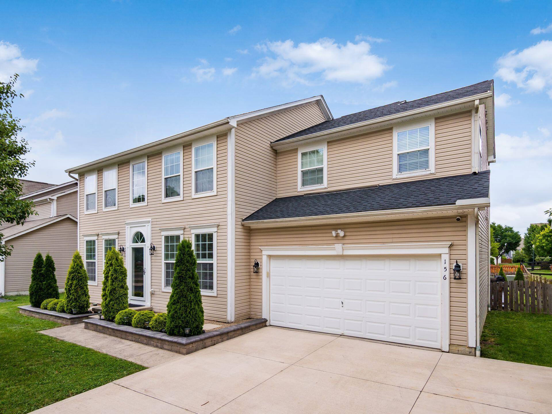 Two-story beige home with a double garage, wide driveway, and neatly trimmed front shrubs.